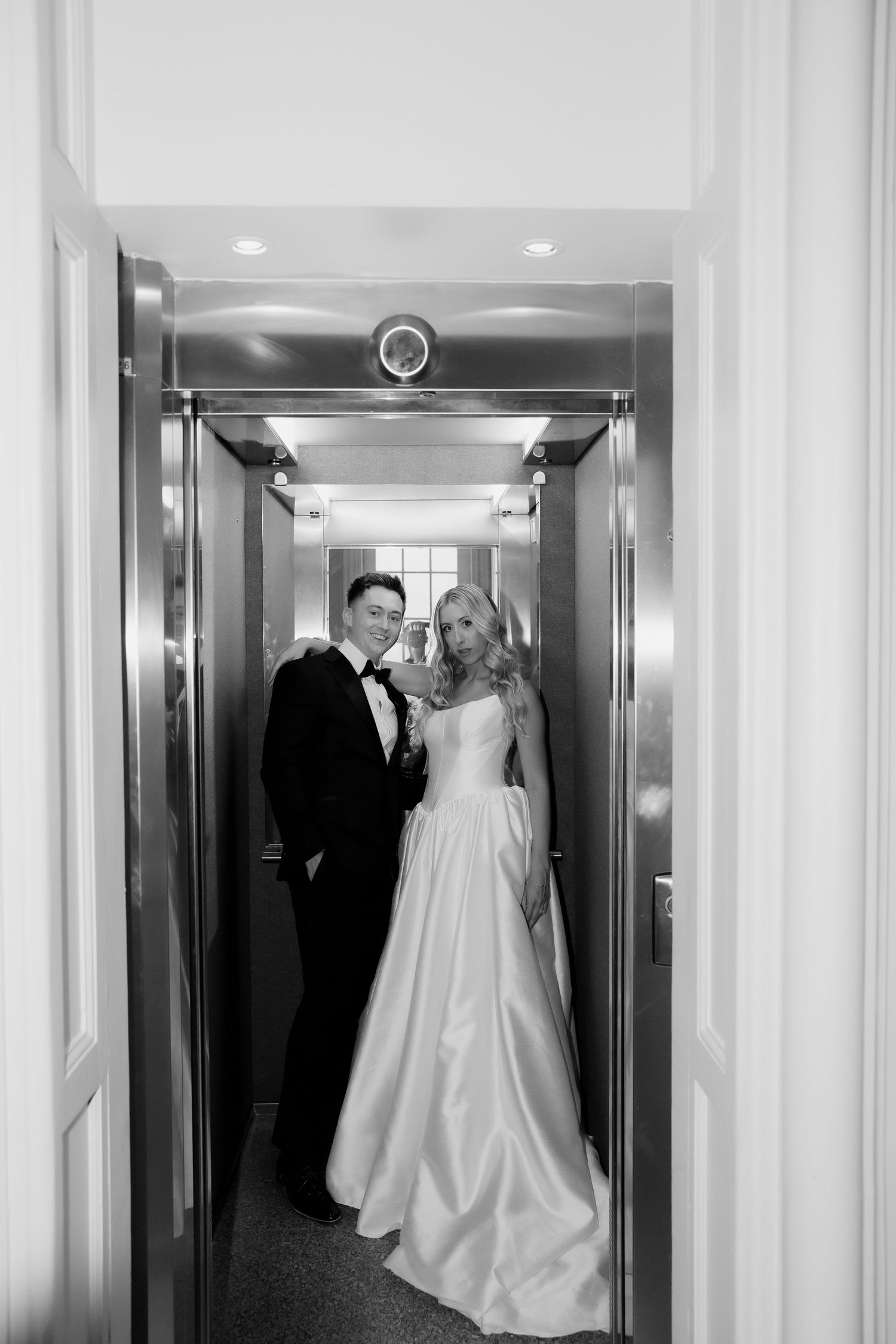 A bride in a white gown and groom in a tuxedo stand together in an elegant elevator on their wedding day.