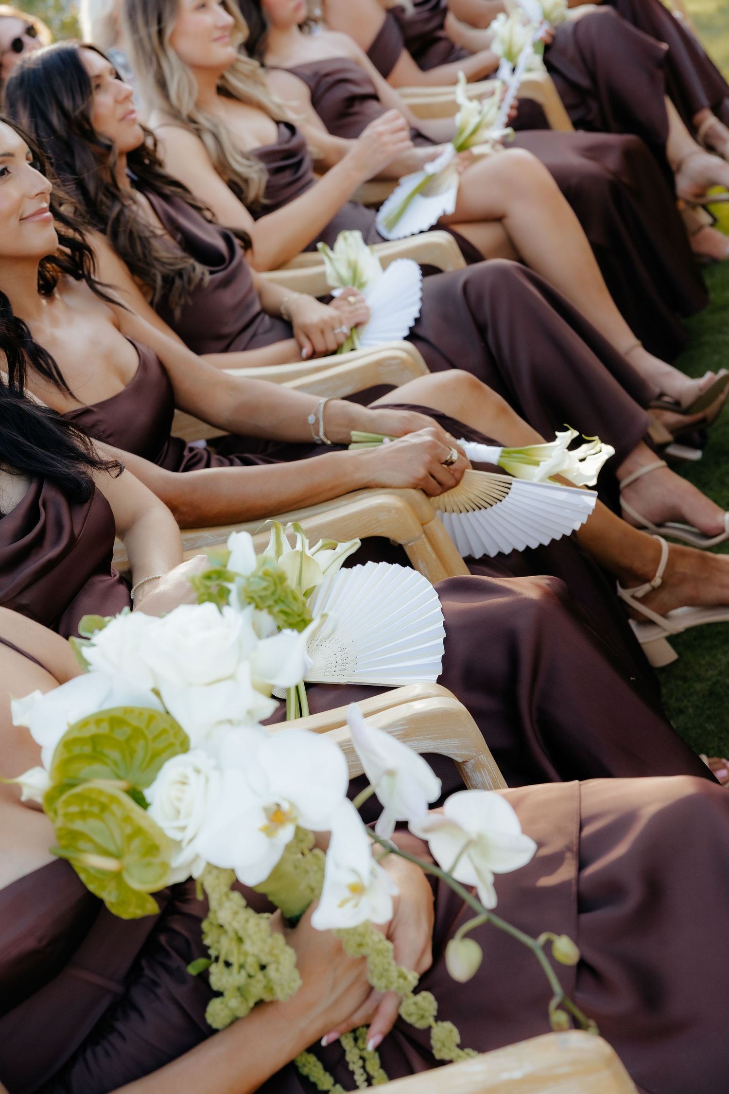 Bridesmaids in brown dresses seated in chairs holding white floral bouquets and paper fans at outdoor wedding ceremony.