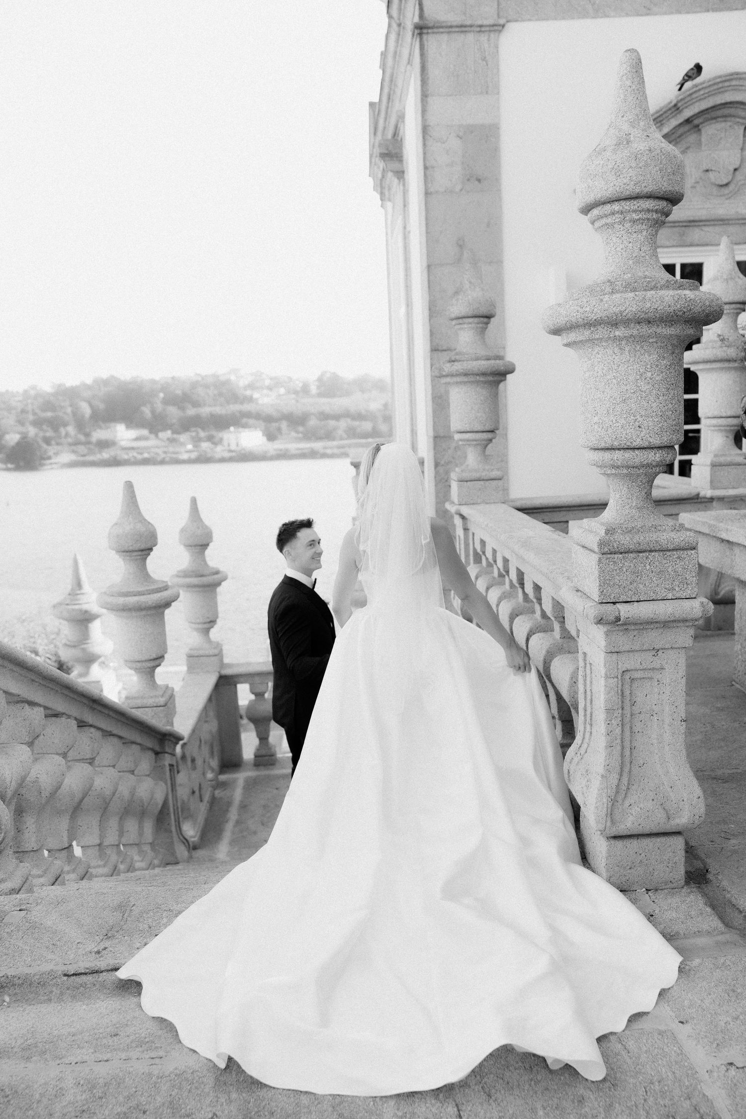 A bride in a flowing white wedding dress stands between elegant stone columns on a balcony in black and white.