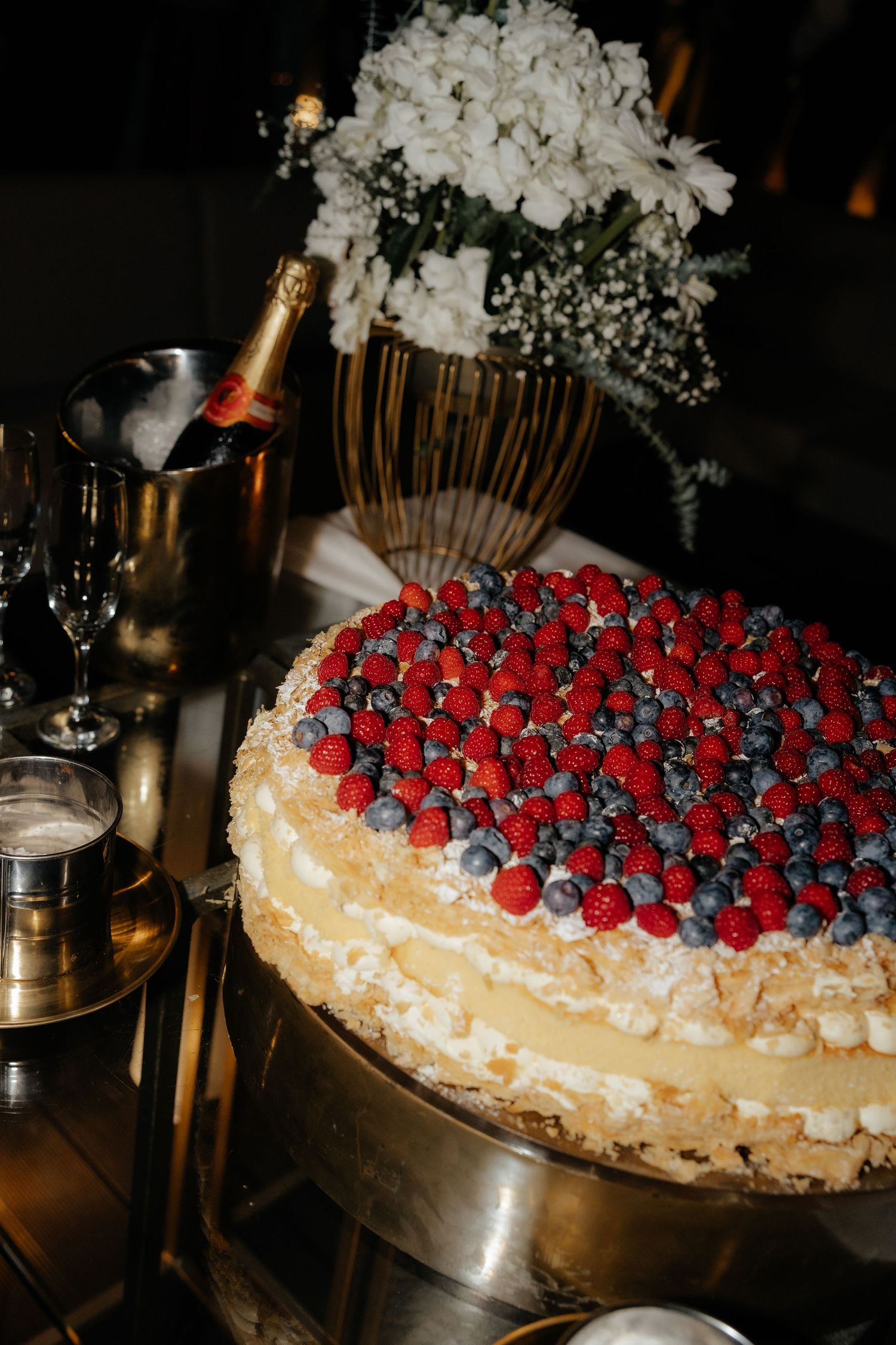 Elegant wedding cake topped with fresh raspberries and blueberries beside champagne and flowers.