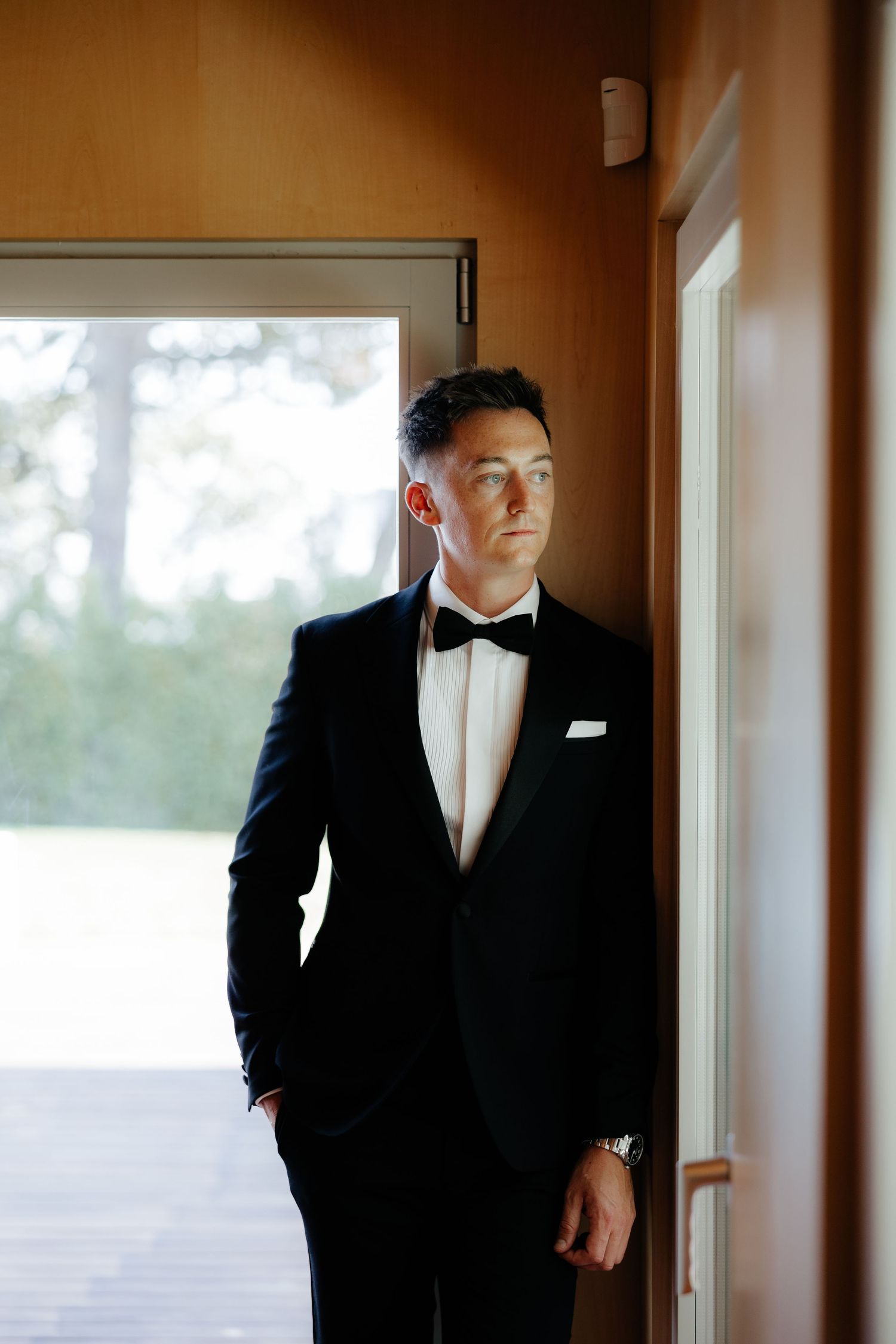 Elegant groom in black tuxedo with bow tie and pocket square leaning against a doorframe in natural light.