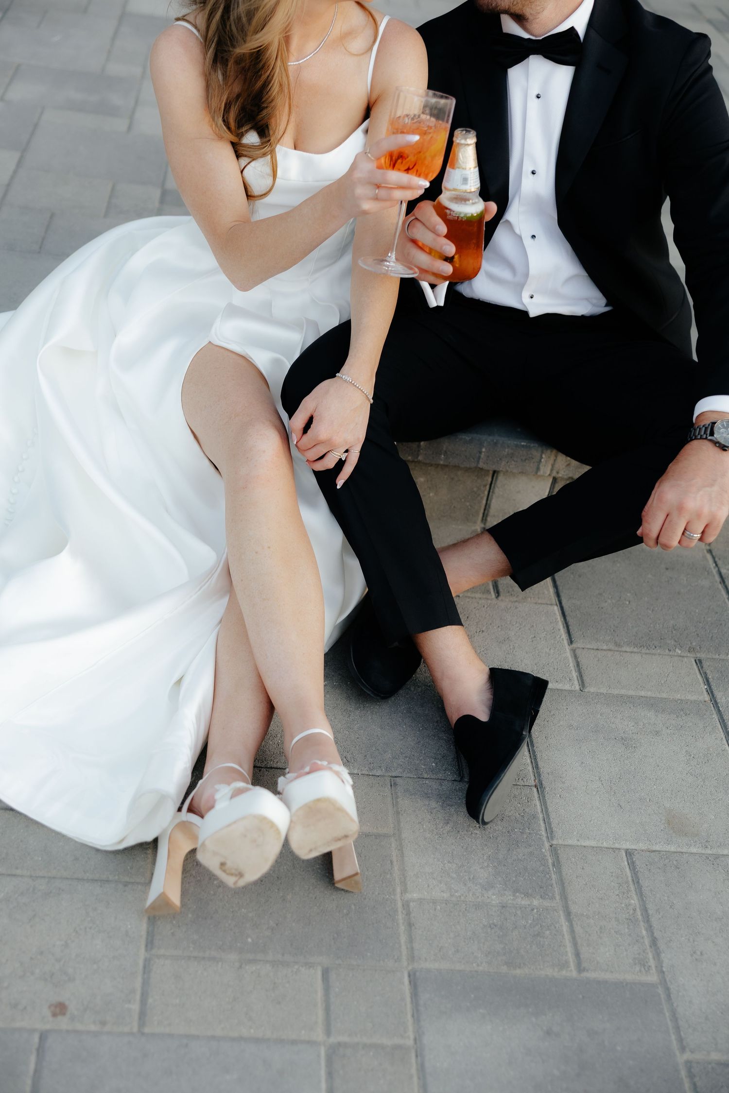 A bride and groom sit together on stone pavers, clinking glasses of orange cocktails at their wedding.