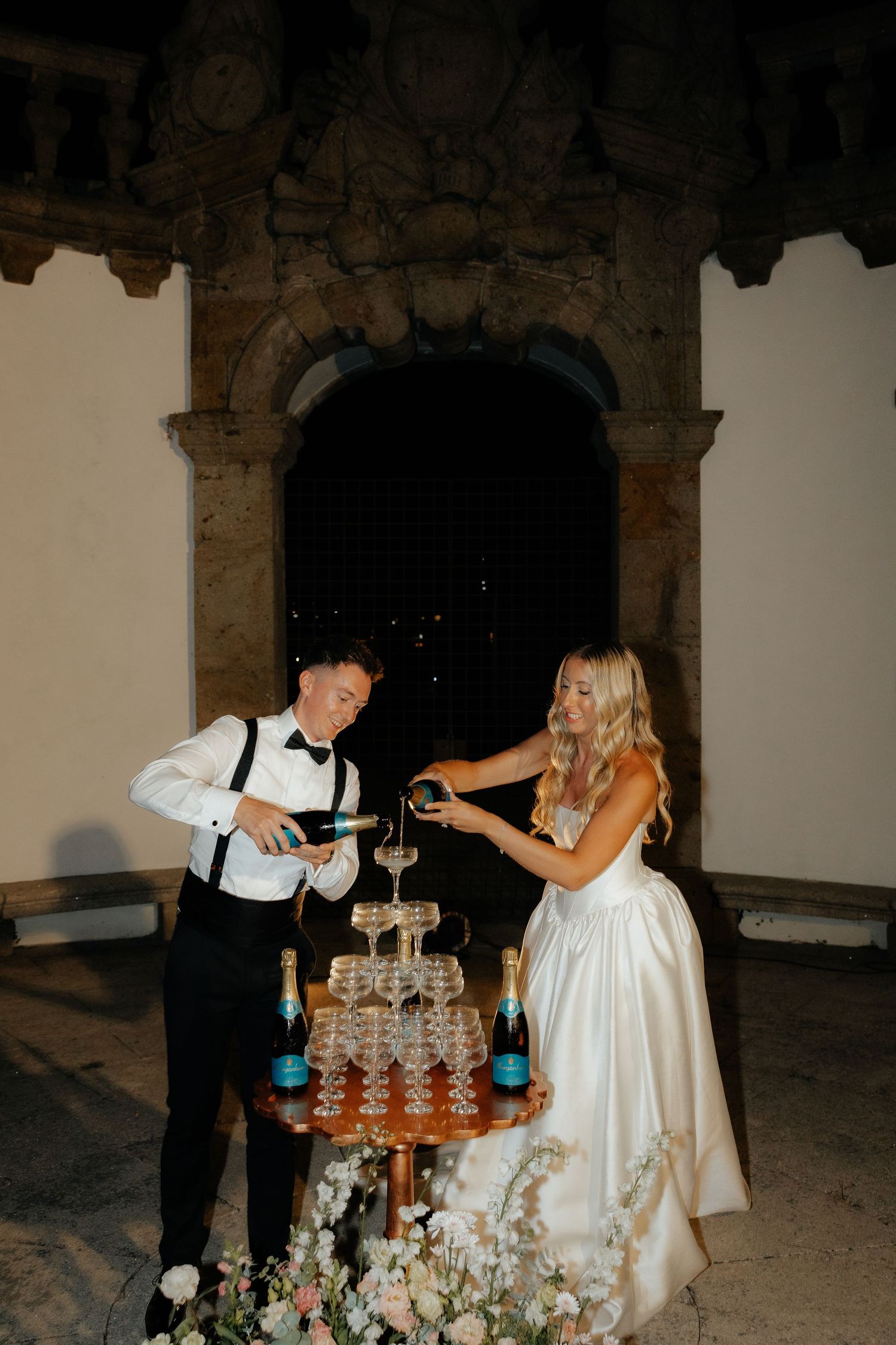 A couple pours champagne into a tower of glasses at an elegant wedding reception in a rustic stone venue.