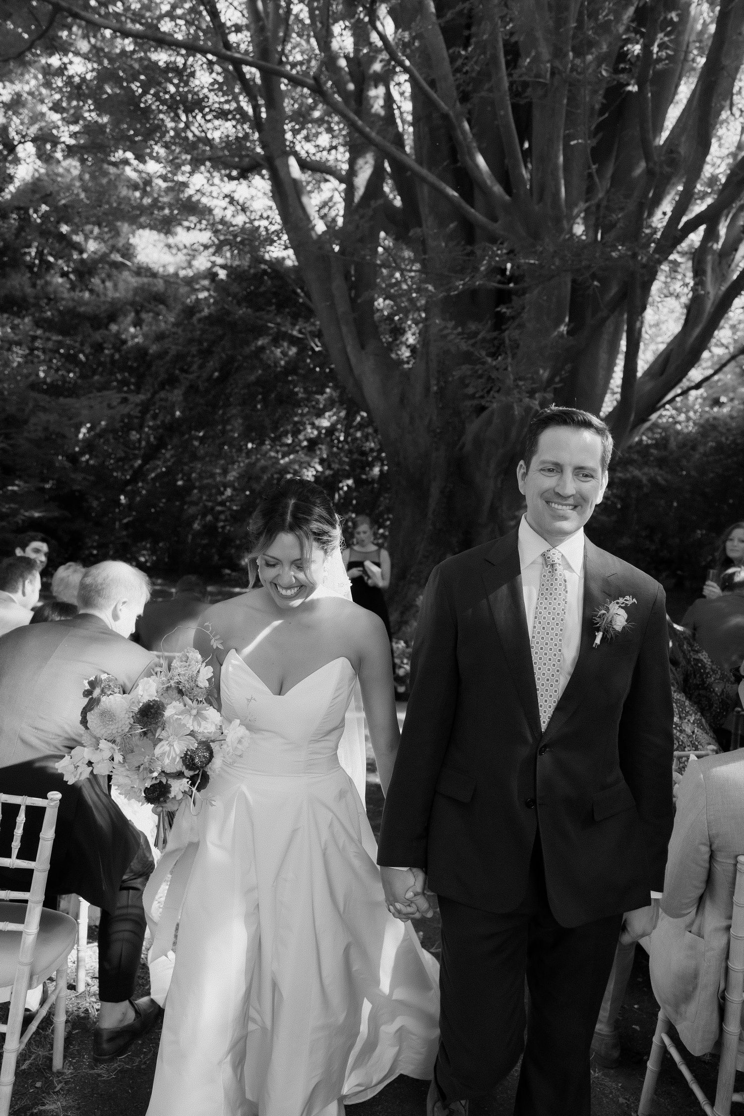 Outdoor wedding ceremony under trees with white chairs and formal attire.