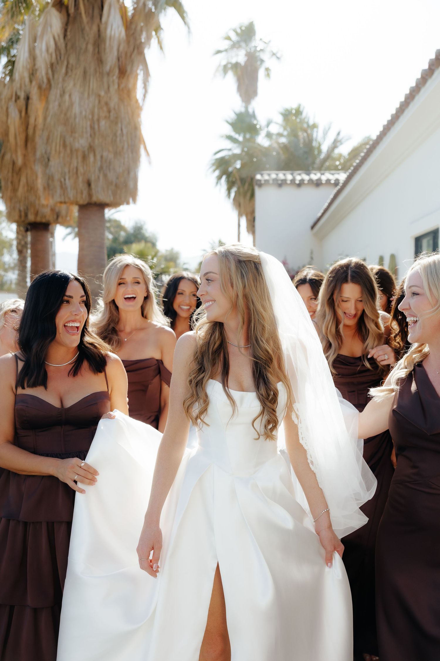 Bride in white gown walking with bridesmaids in dark brown dresses near palm trees.