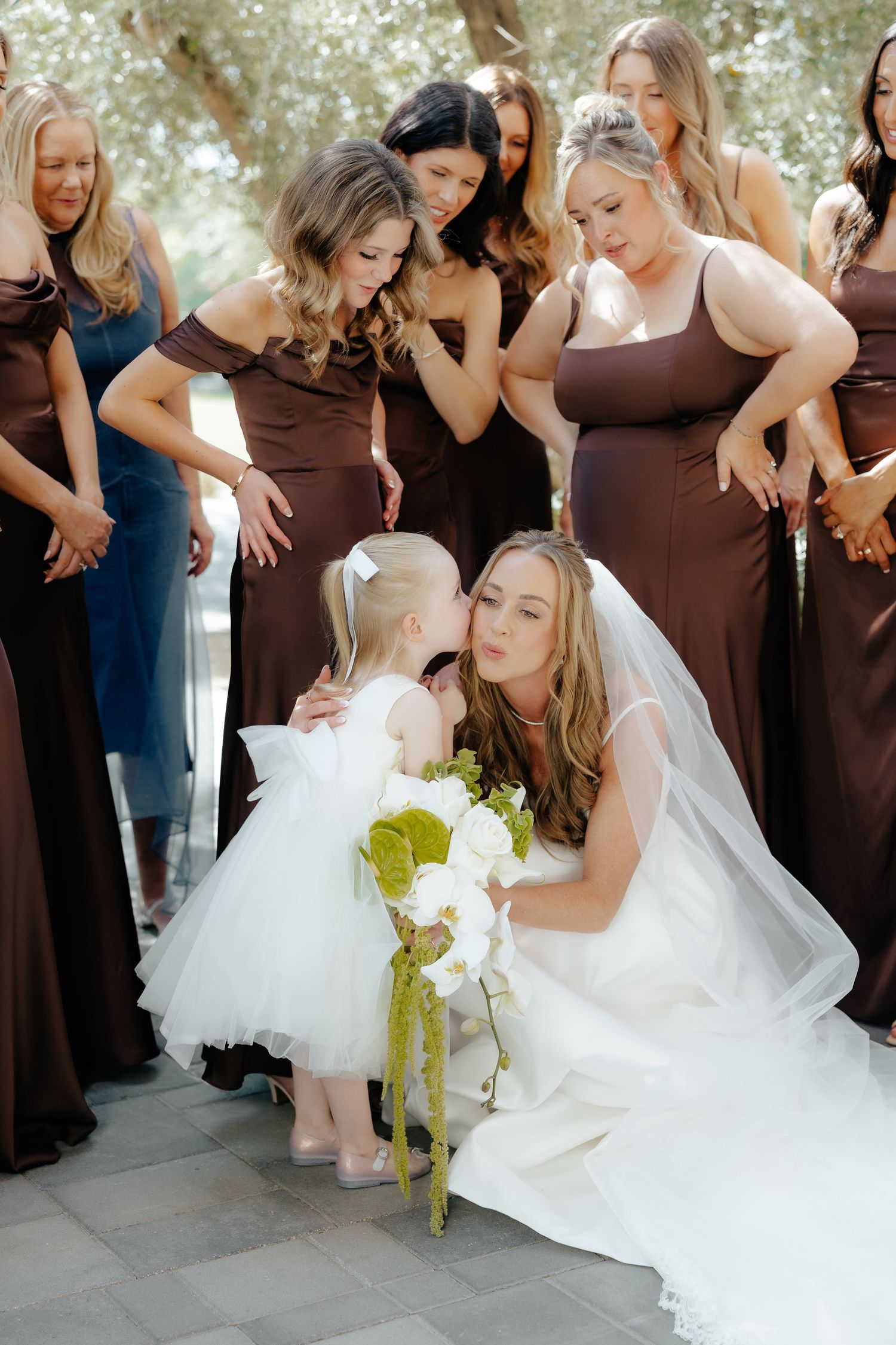 Bride in white gown crouching to kiss a flower girl surrounded by brown-gowned bridesmaids.