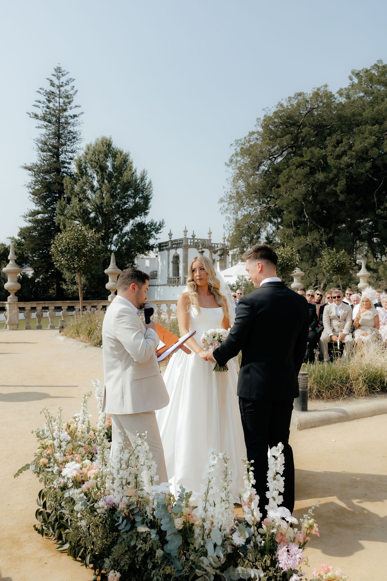 An outdoor wedding ceremony takes place in a garden setting with floral arrangements and a classical architectural backdrop.