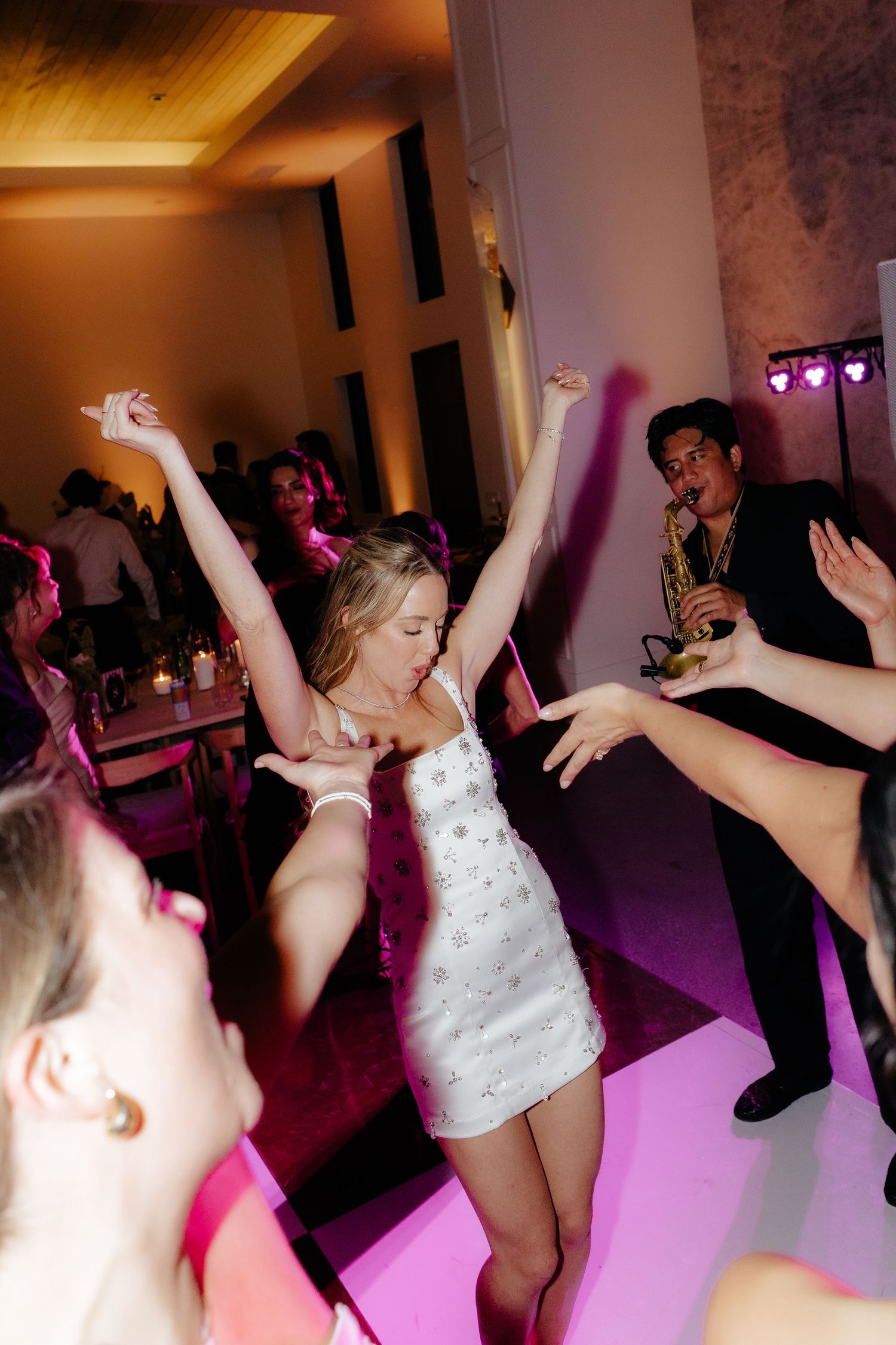 Bride in white mini dress dancing with arms raised at wedding reception with saxophonist performing on stage.