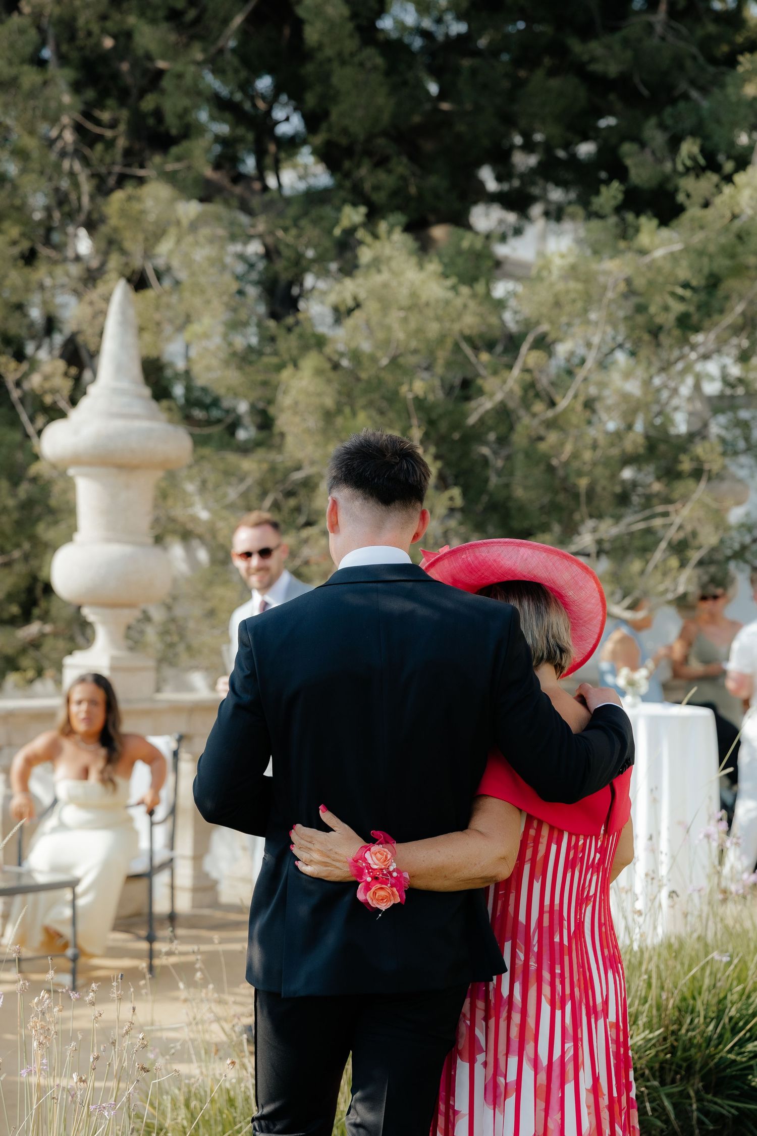 People in formal attire walk through a cemetery with white crosses and ornate stone monuments in the background.