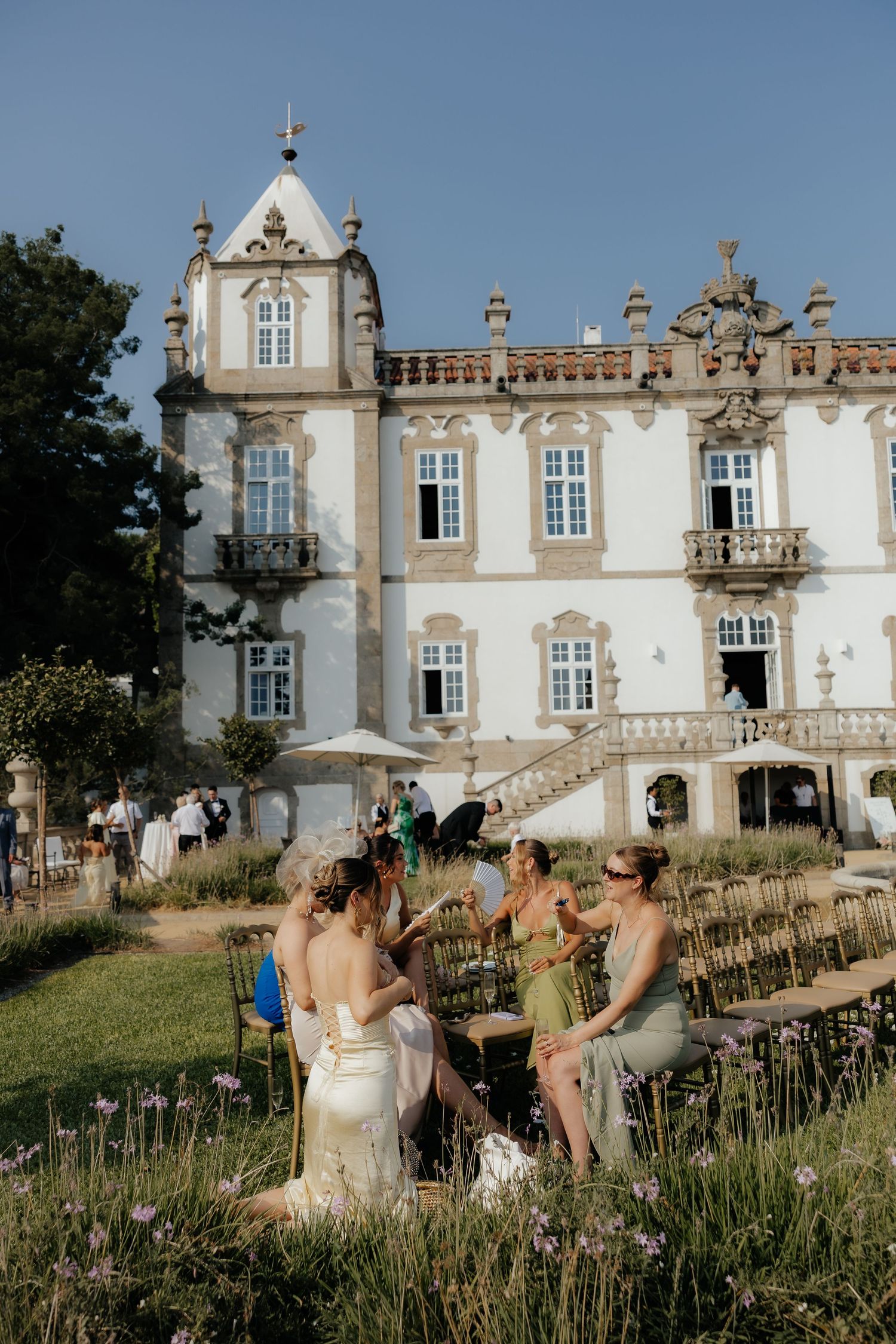 People enjoying drinks on a lawn outside an elegant white historic mansion with ornate architecture on a sunny day.