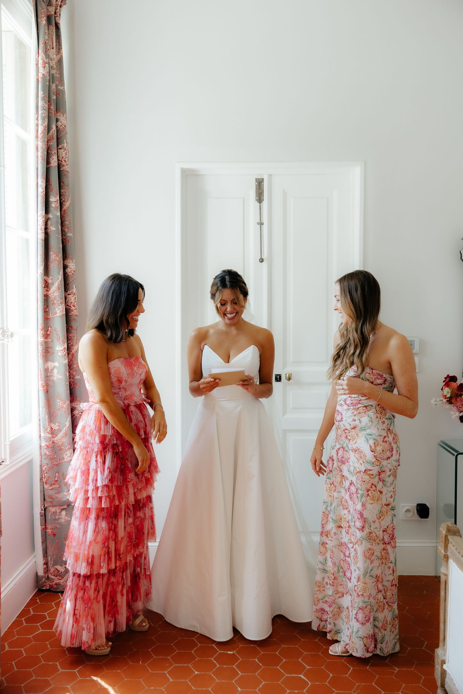 Bridesmaids in coral floral dresses helping with wedding preparations in a bright room.