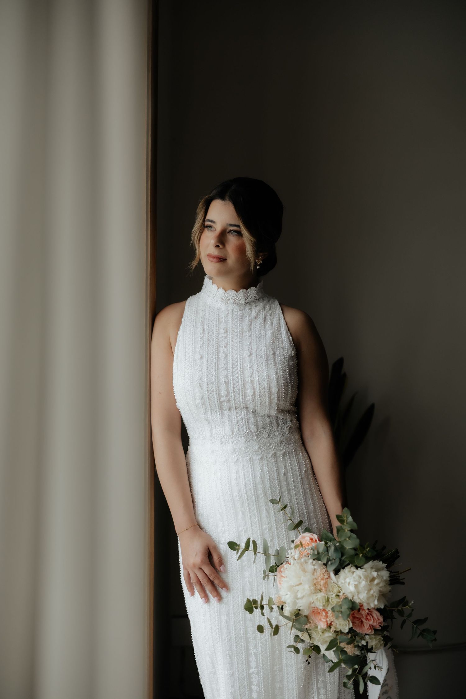 Elegant bride in a white sleeveless lace gown holding a bouquet while standing by a window.