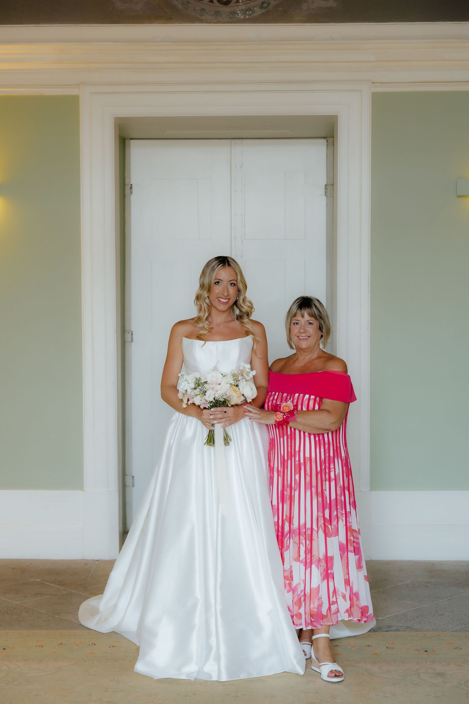 Bride in white strapless ball gown poses with woman in pink floral dress holding bouquet.