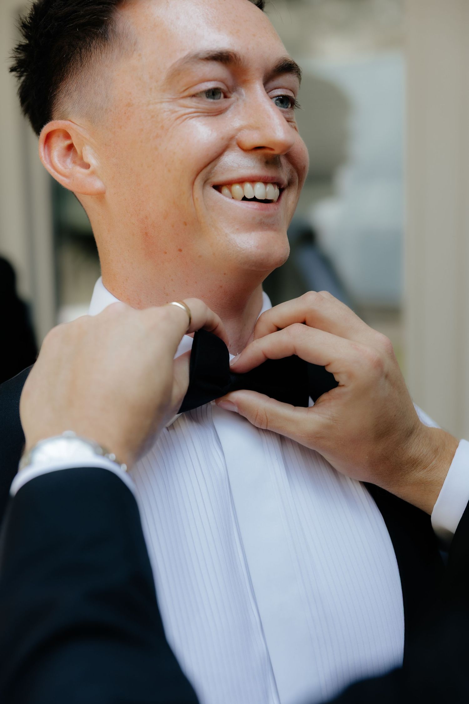 Smiling groom in black tuxedo having his bow tie adjusted before his wedding ceremony.