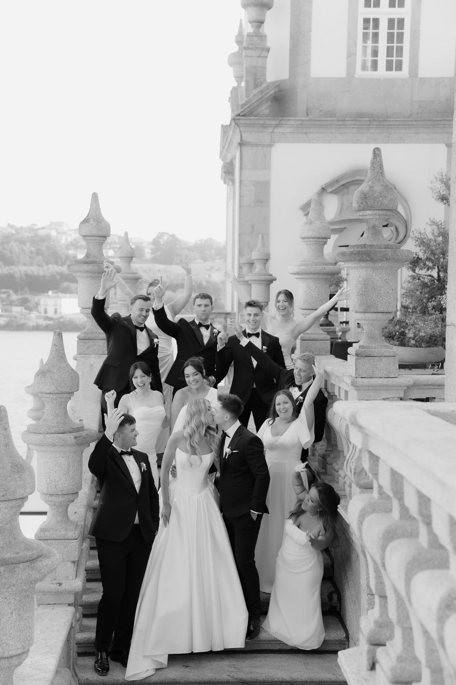 Wedding party celebrating on an elegant stone balcony overlooking water, raising glasses in black and white photo.