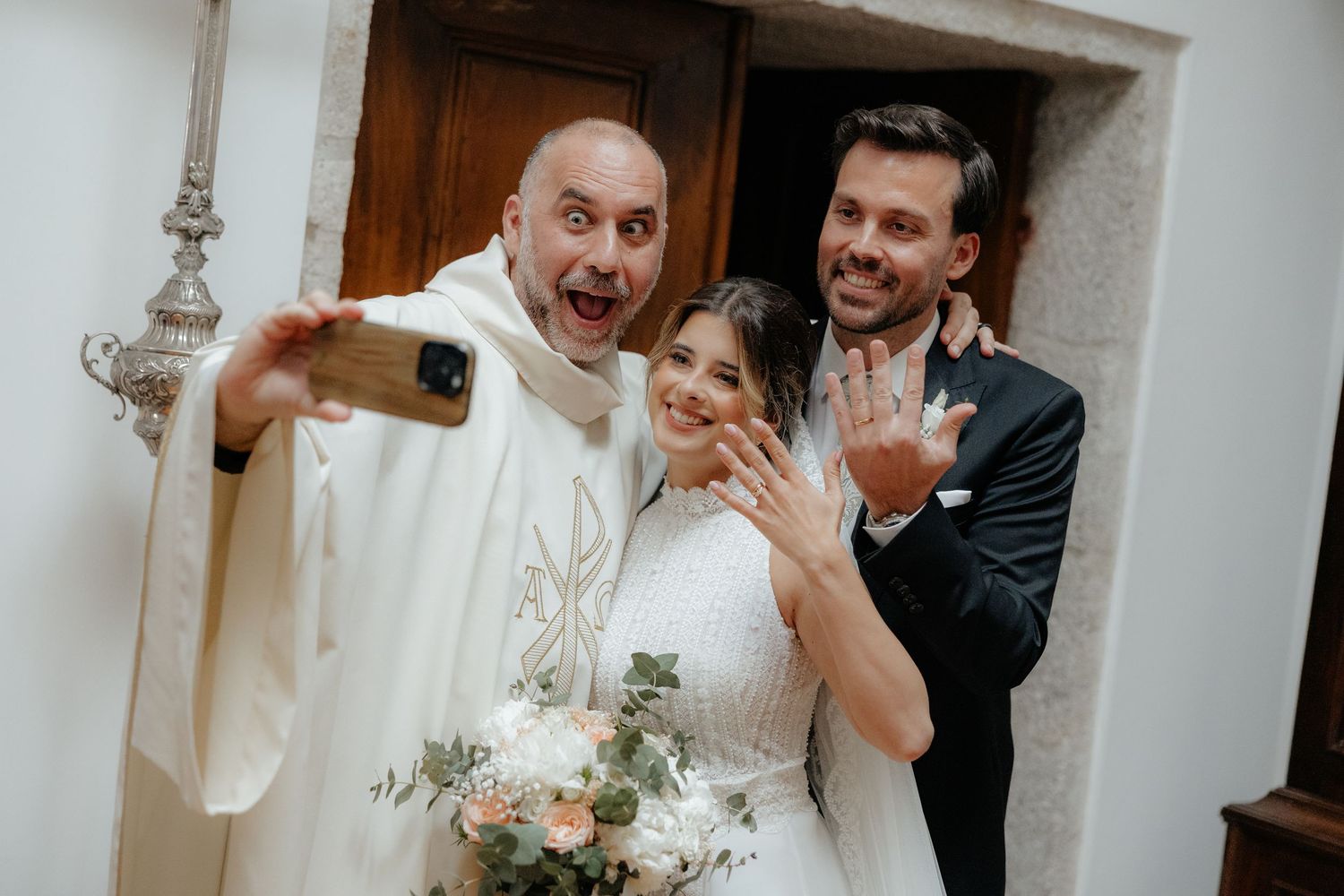 Excited priest takes selfie with newlyweds showing their wedding rings after the ceremony.