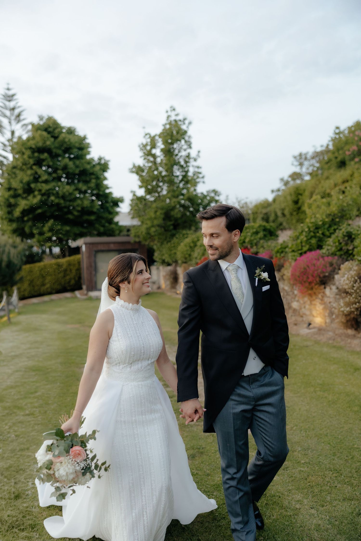 Bride and groom walk hand in hand through lush garden, bride holding bouquet in elegant white dress.