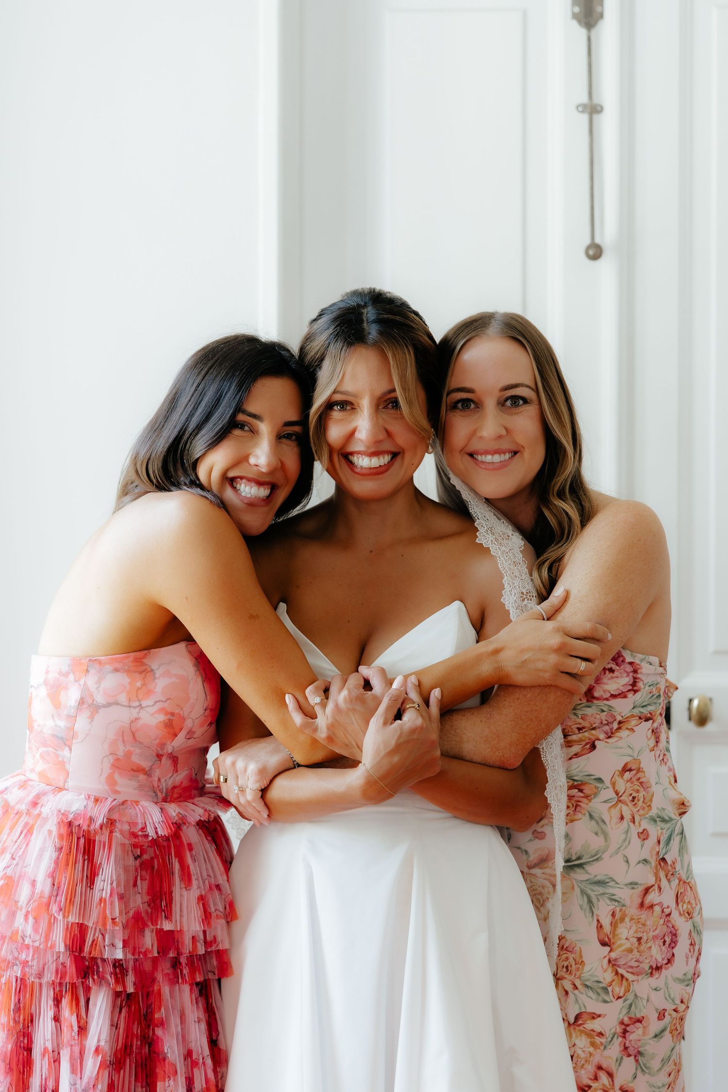 Bridesmaids in pink floral dresses sharing a joyful group hug.
