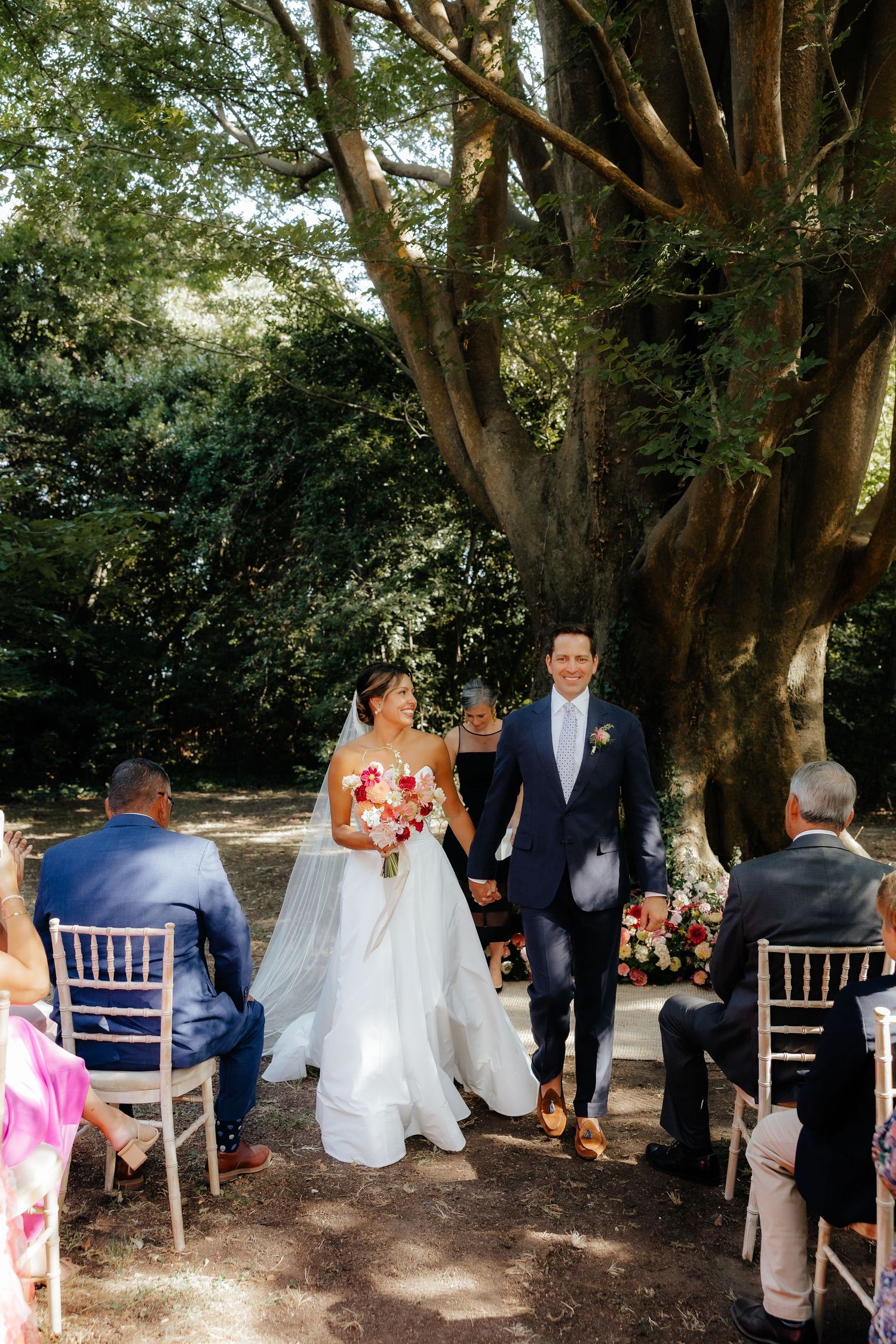 A garden wedding reception with white chairs arranged under large shady trees.