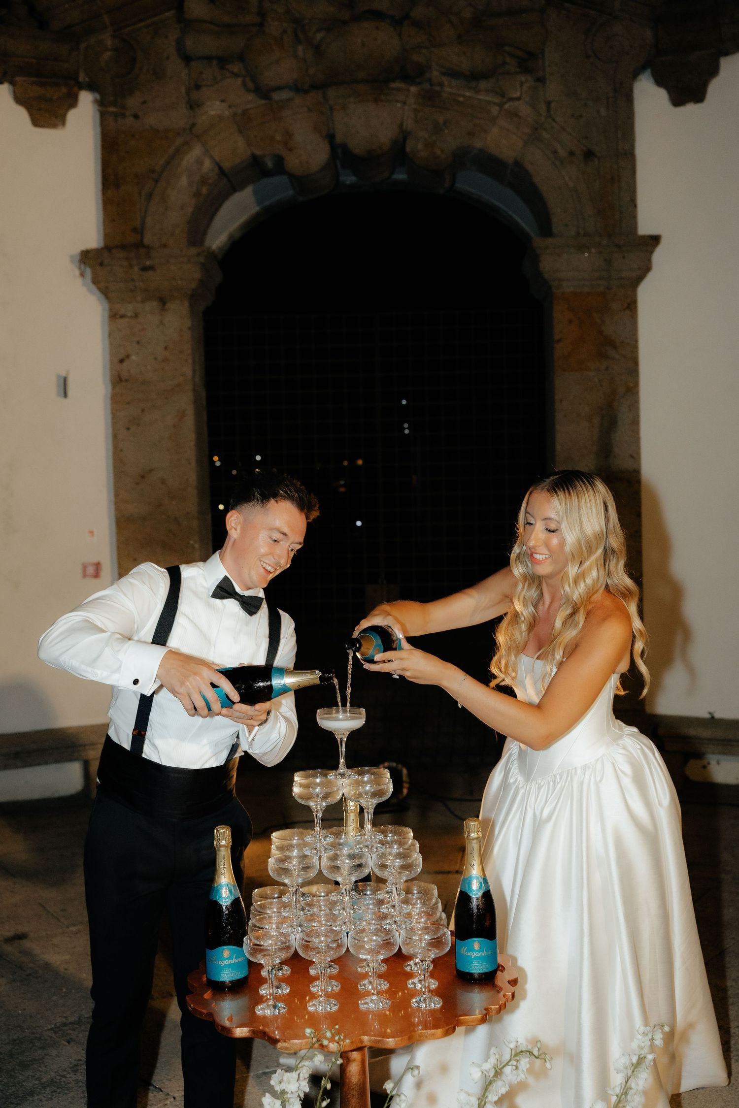 A couple pours champagne into a tower of glasses during an elegant wedding reception celebration.
