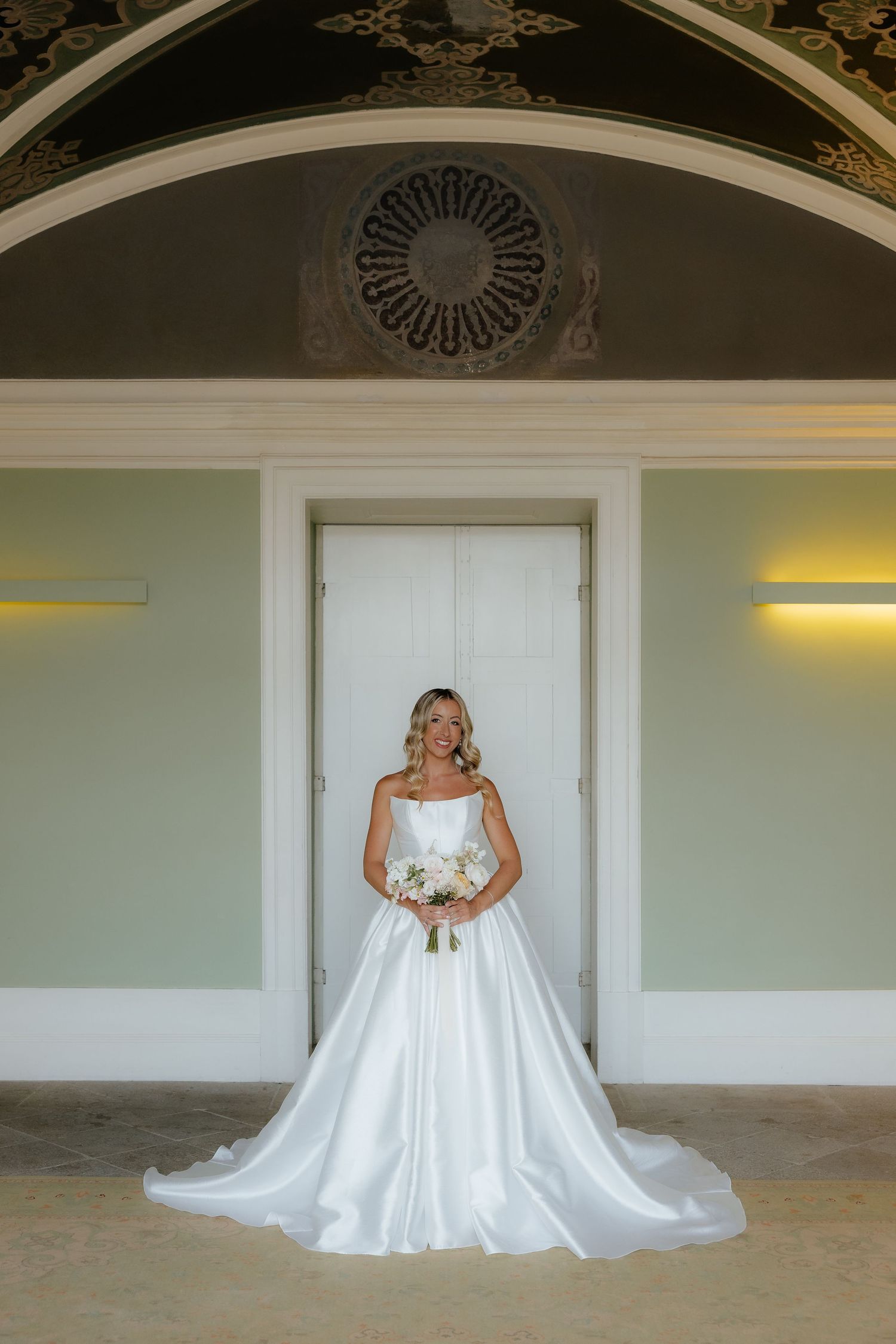 A bride in a flowing white satin wedding gown stands elegantly against a light-colored wall in a grand hallway.