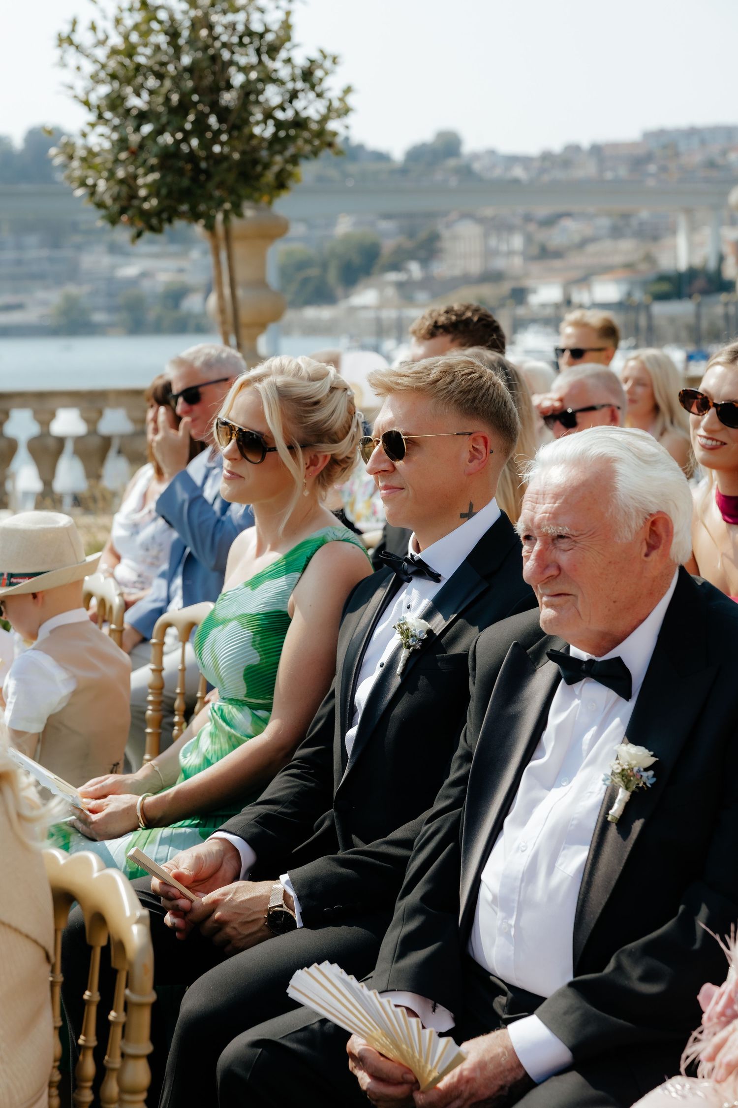 Wedding guests seated in formal attire watch an outdoor ceremony on a sunny day by the water.
