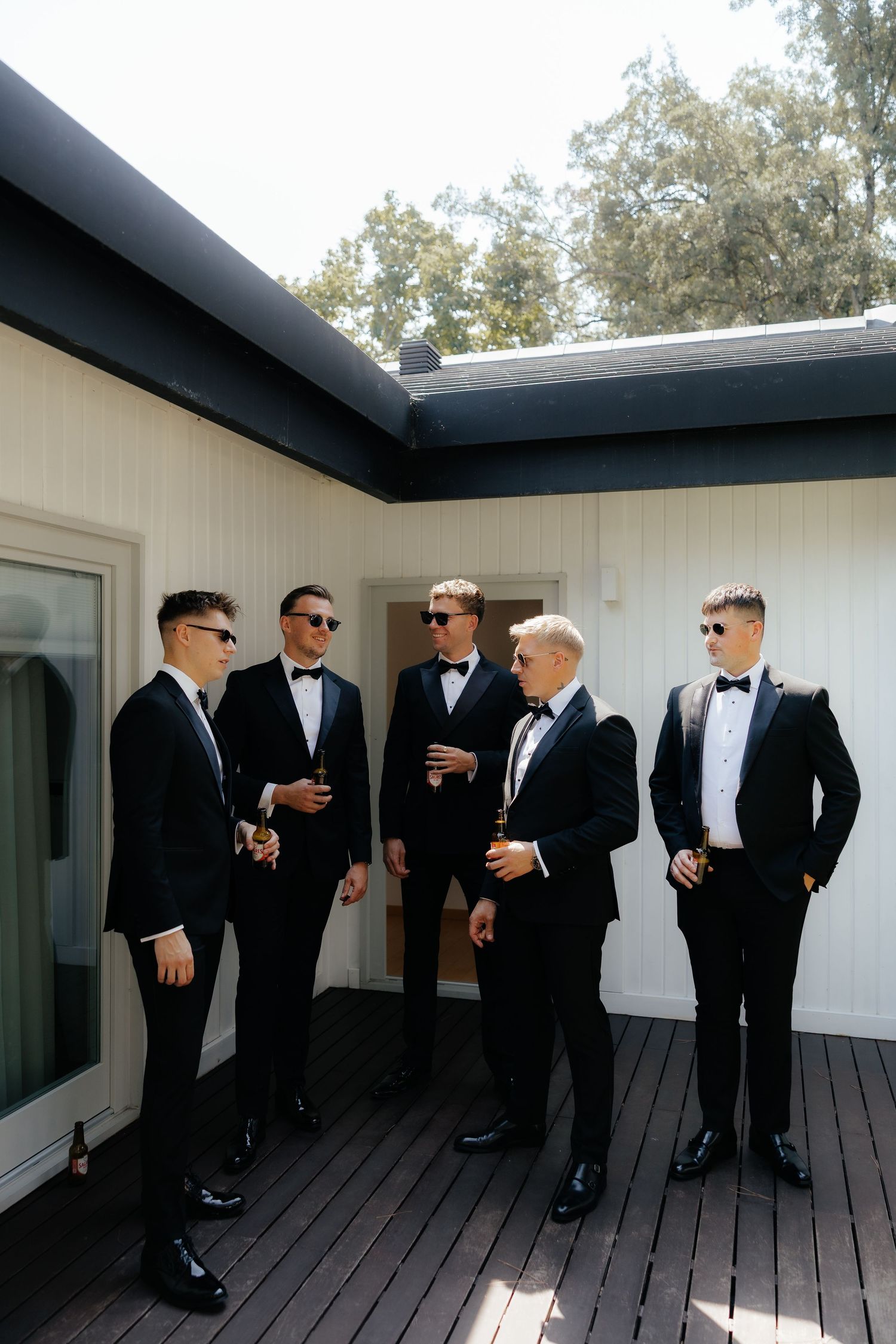 A group of groomsmen in black tuxedos and bow ties gather on a wooden deck outside a white building.