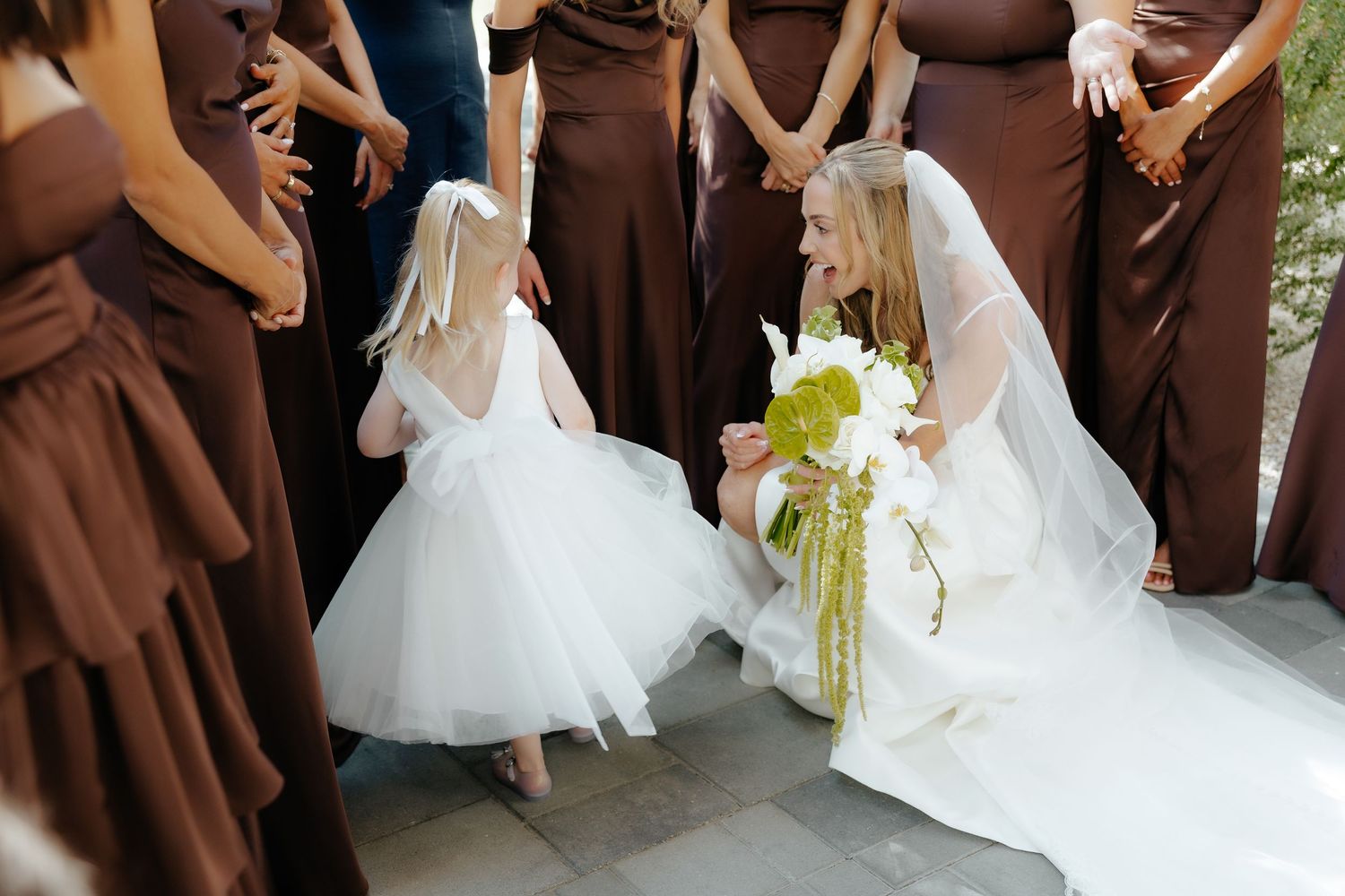 Bride kneeling to greet flower girl in white dress among brown-gowned bridesmaids outdoors.