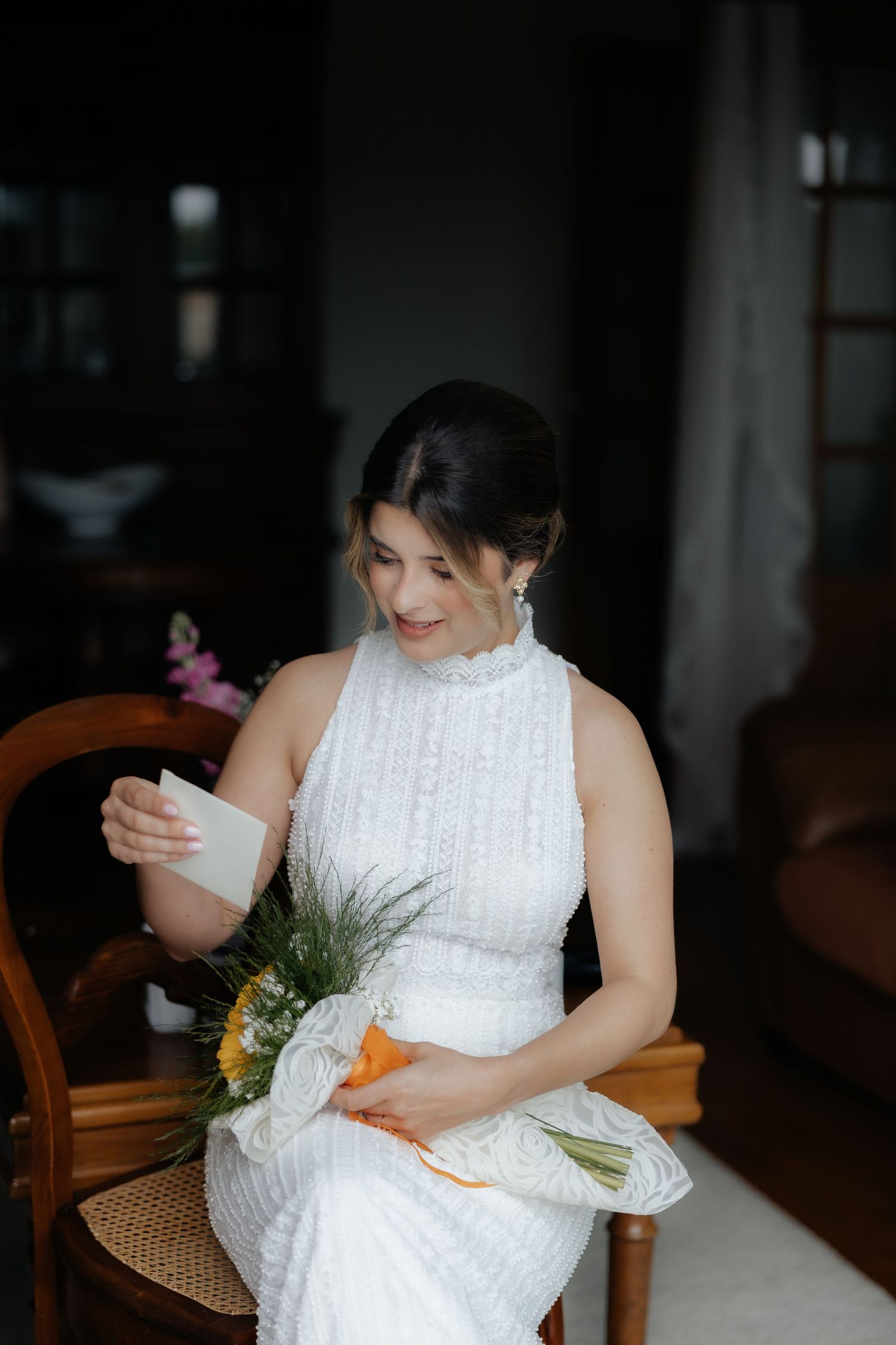 Smiling bride in a white lace dress sitting on a chair reading a letter while holding a bouquet.