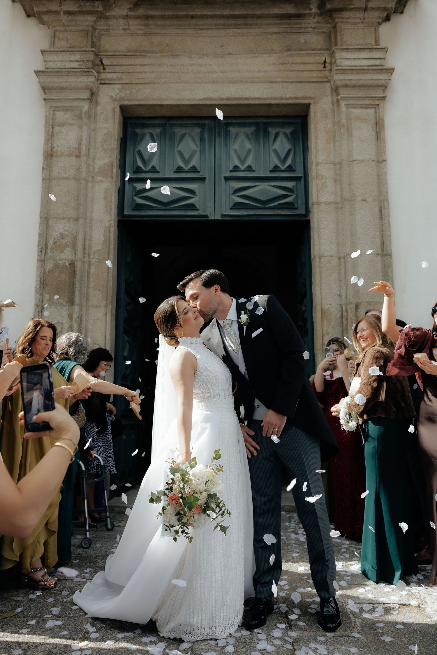 Newlyweds share romantic kiss outside church entrance surrounded by guests tossing white confetti petals.
