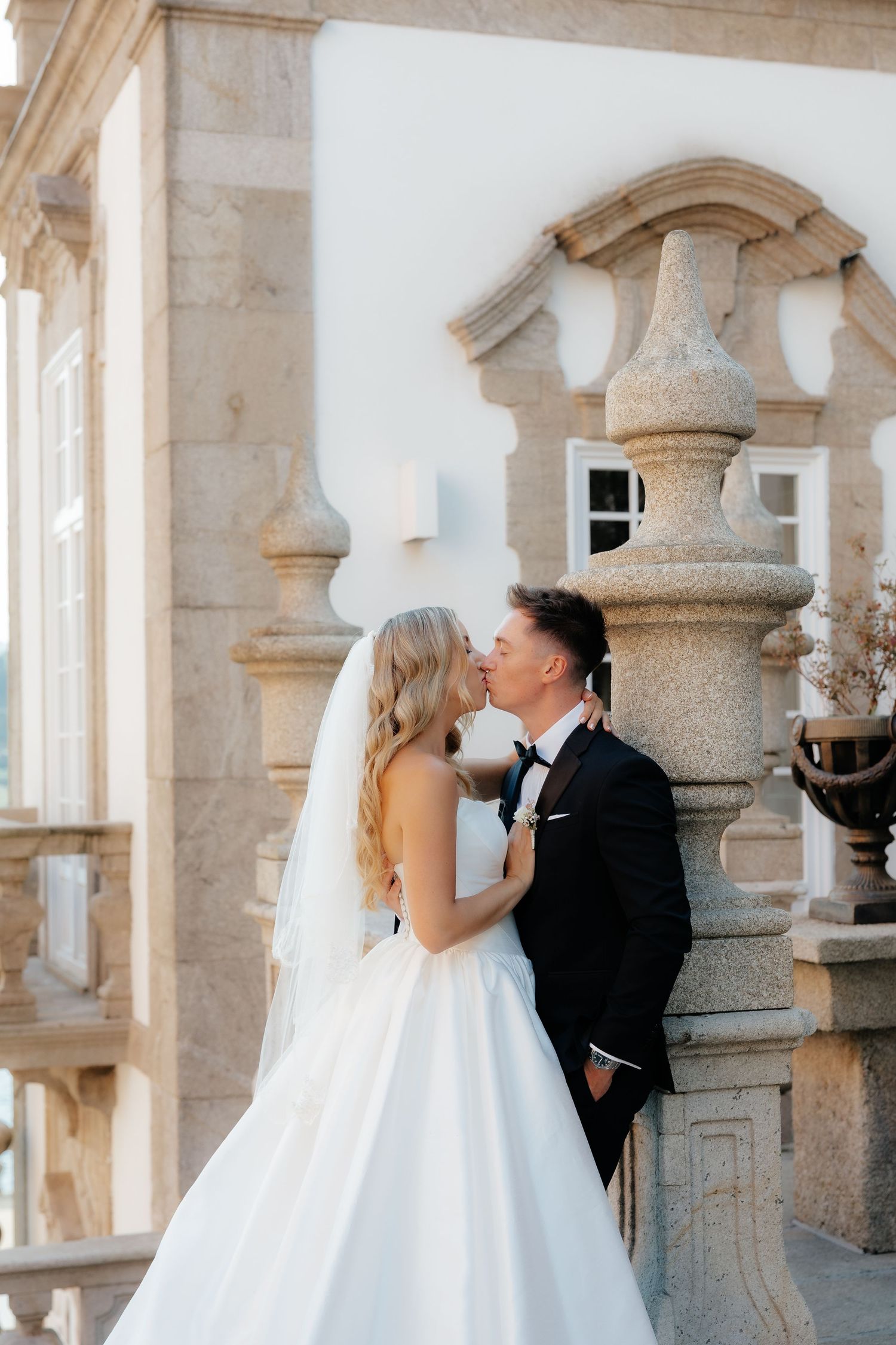 Bride and groom share a kiss beside decorative stone pillar at elegant venue in warm sunlight.