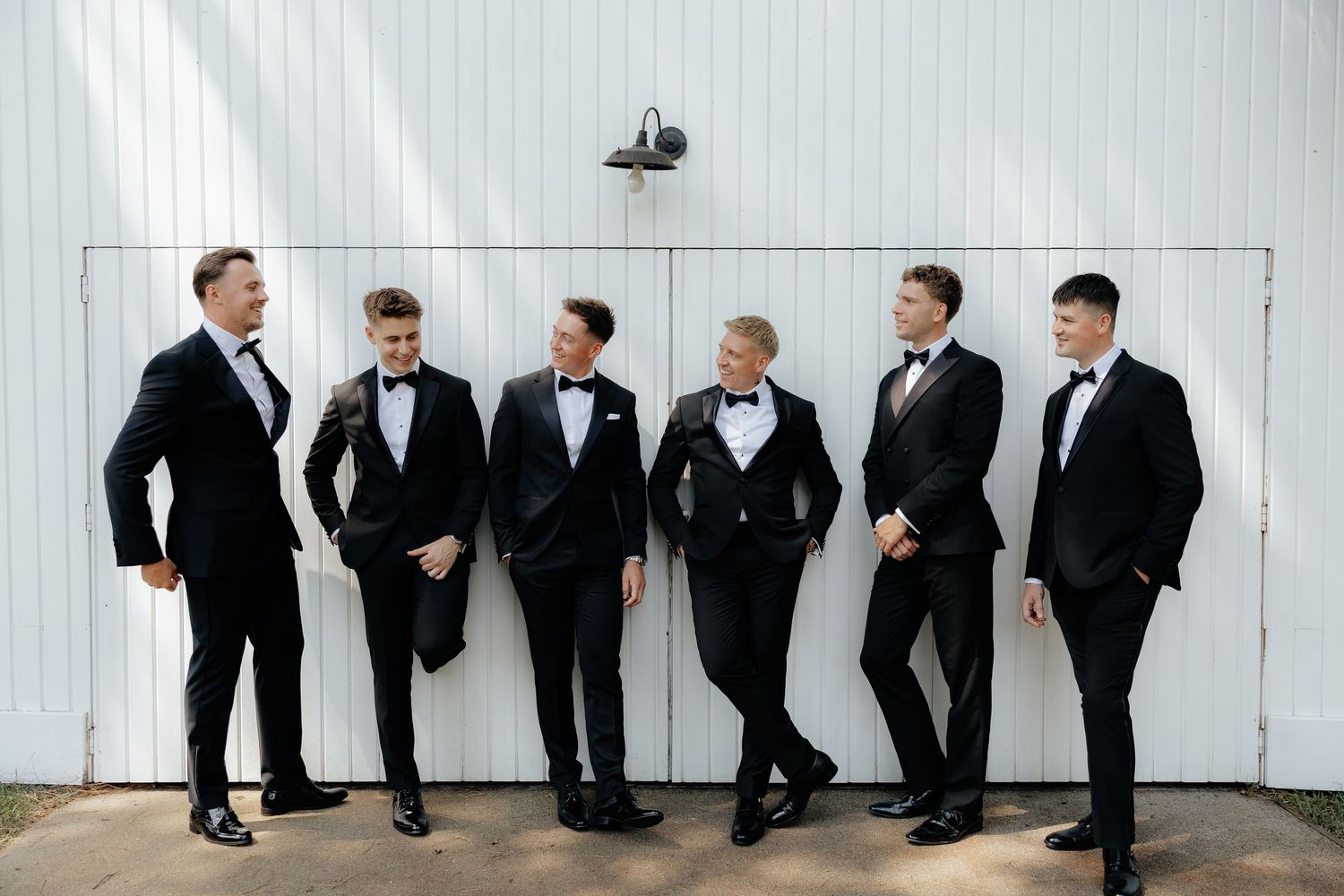 Six groomsmen in black tuxedos posing casually against a white barn wall outdoors.