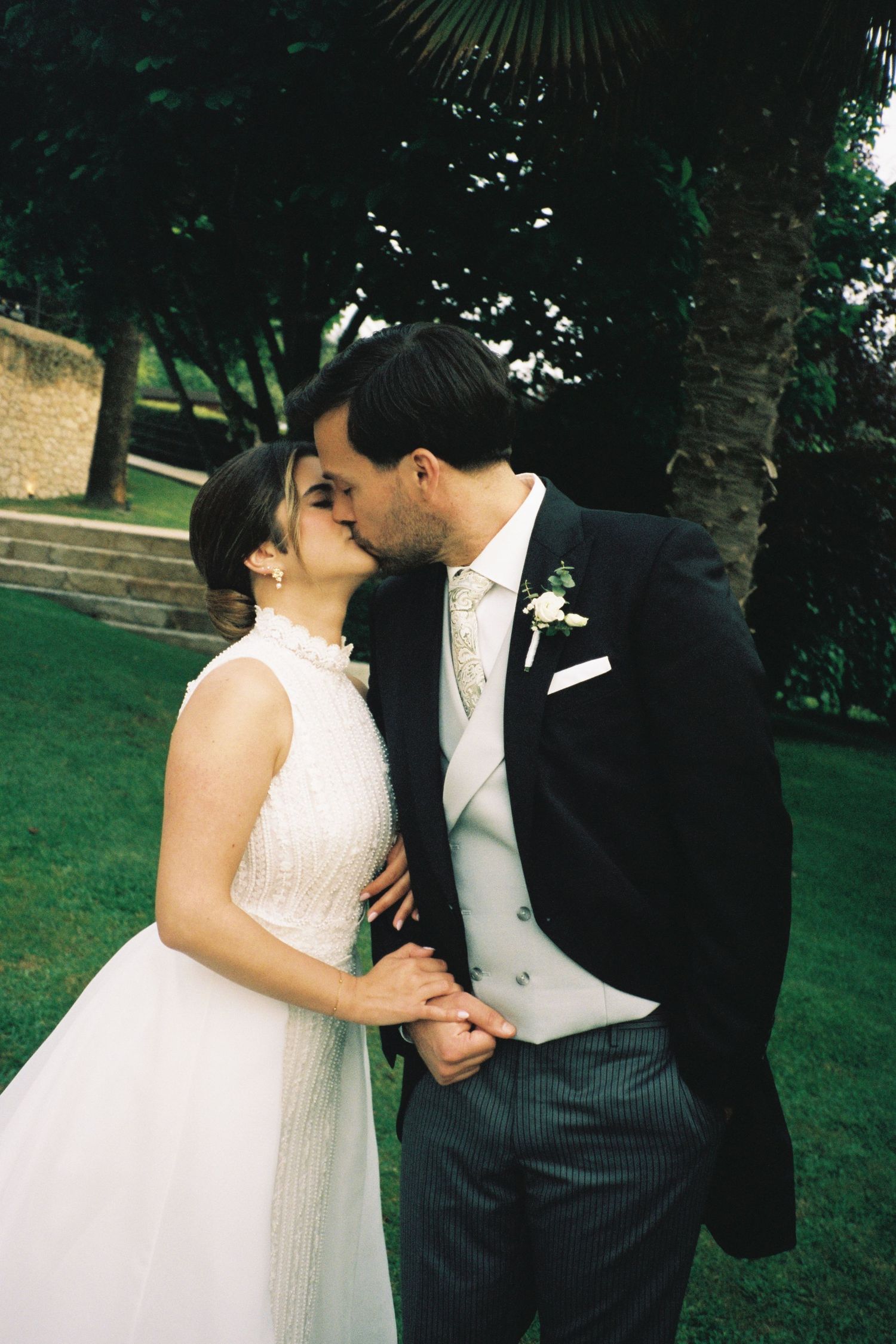 Bride and groom share a tender kiss while holding hands in a lush green garden setting.