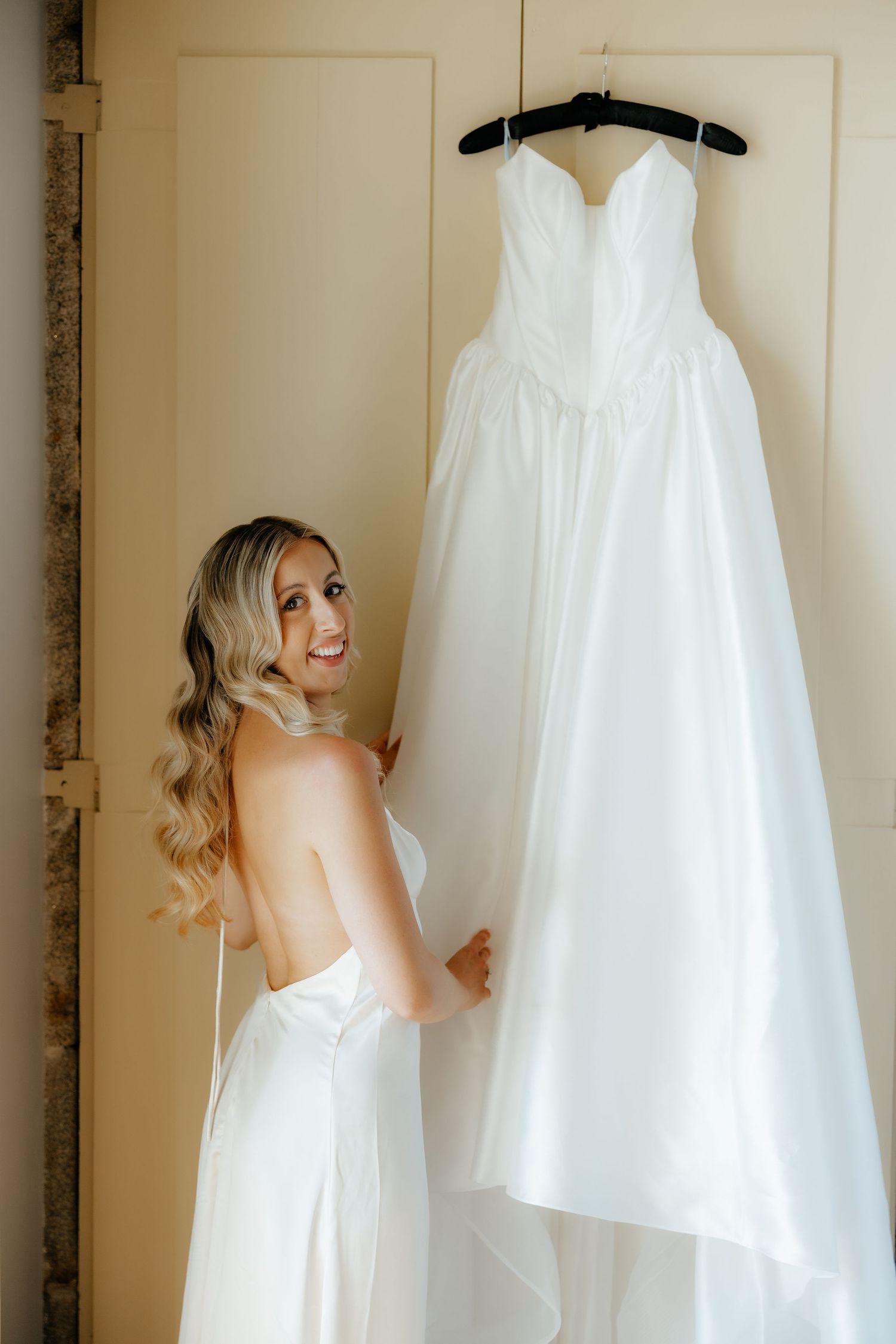 Radiant blonde bride smiles back at camera while touching her elegant strapless wedding dress.