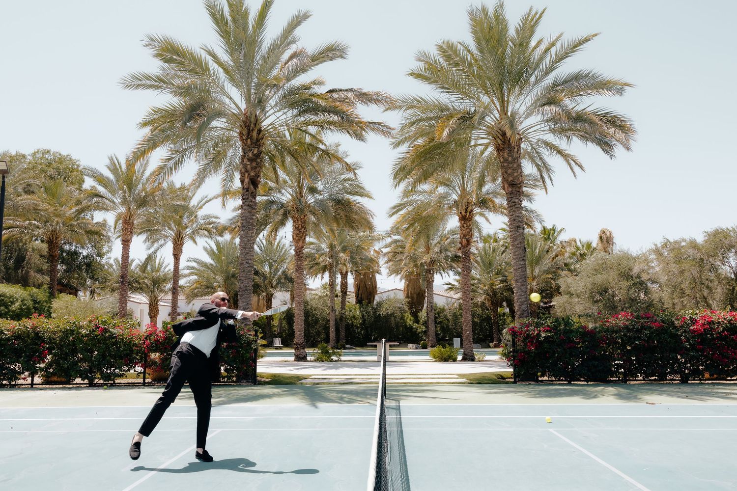 Groom serving tennis ball in tuxedo on sunny court lined with tall palm trees.