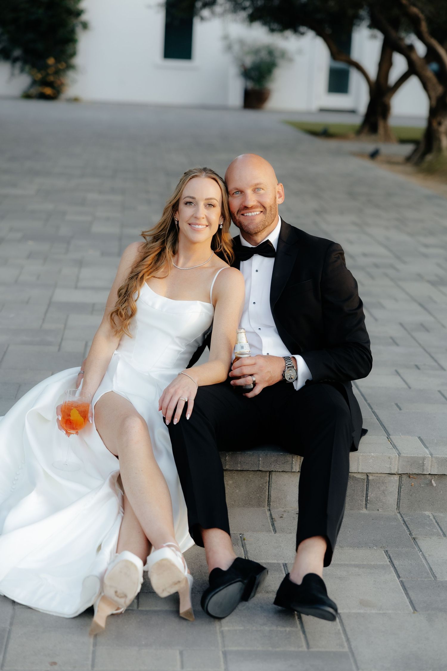 Bride and groom sitting on stone steps smiling, bride in white spaghetti strap gown, groom in black tuxedo.