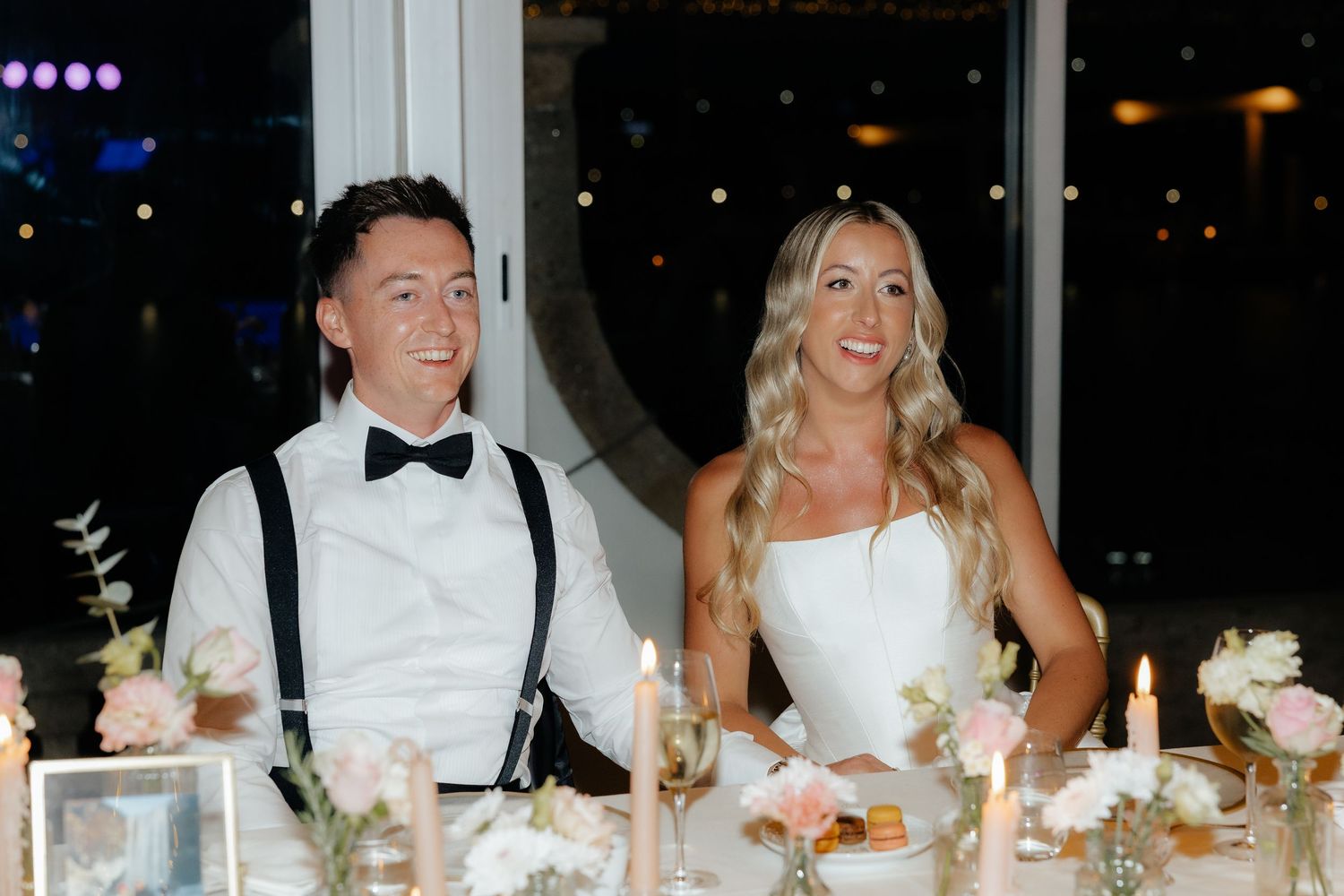 Wedding reception guests seated at a long dining table with floral centerpieces and glowing candlelight.