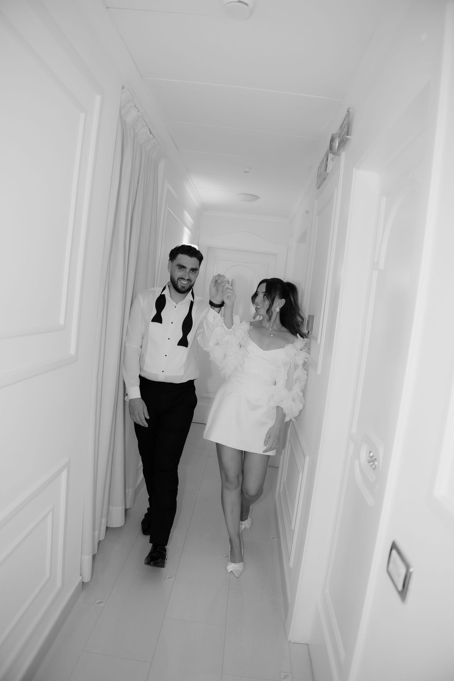 Black and white photo of a couple walking hand in hand down a bright hallway, dressed in wedding attire.
