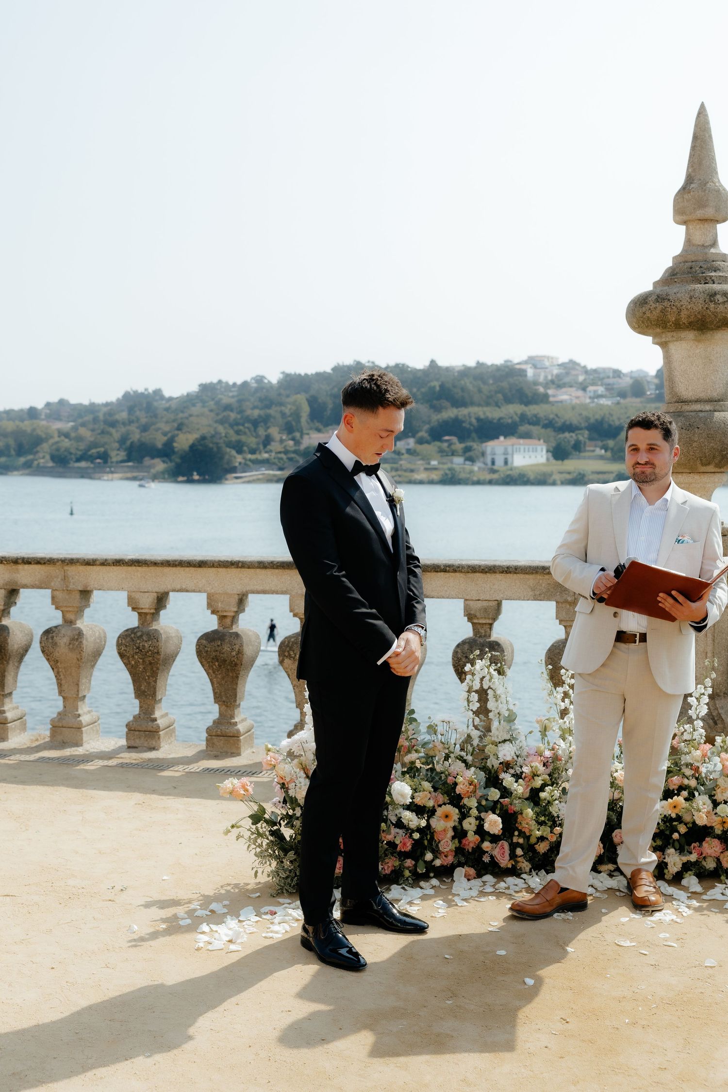 Groom in black tuxedo stands at outdoor altar with officiant beside waterfront balustrade.