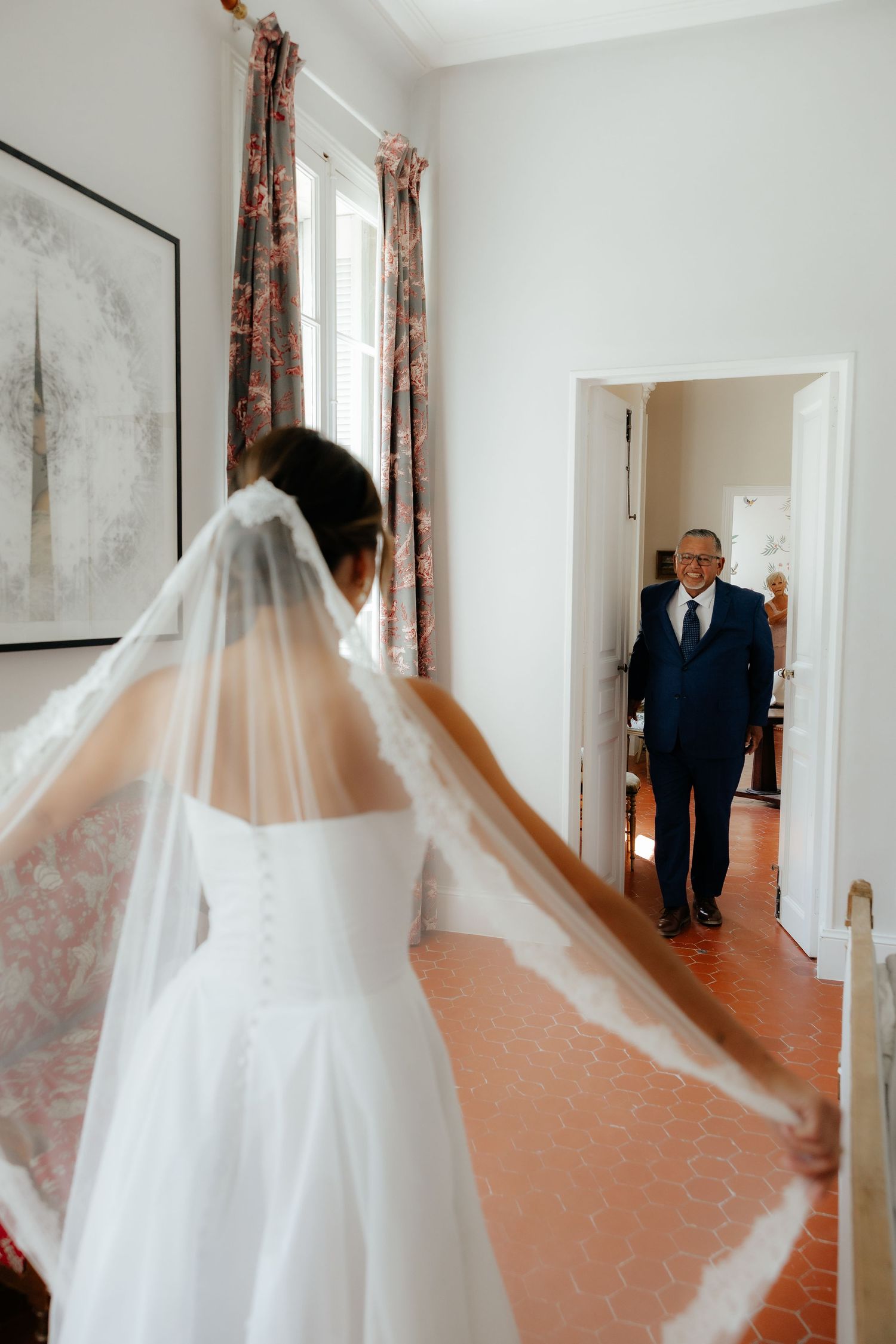 A flowing white wedding dress and veil captured in a hallway with floral curtains.