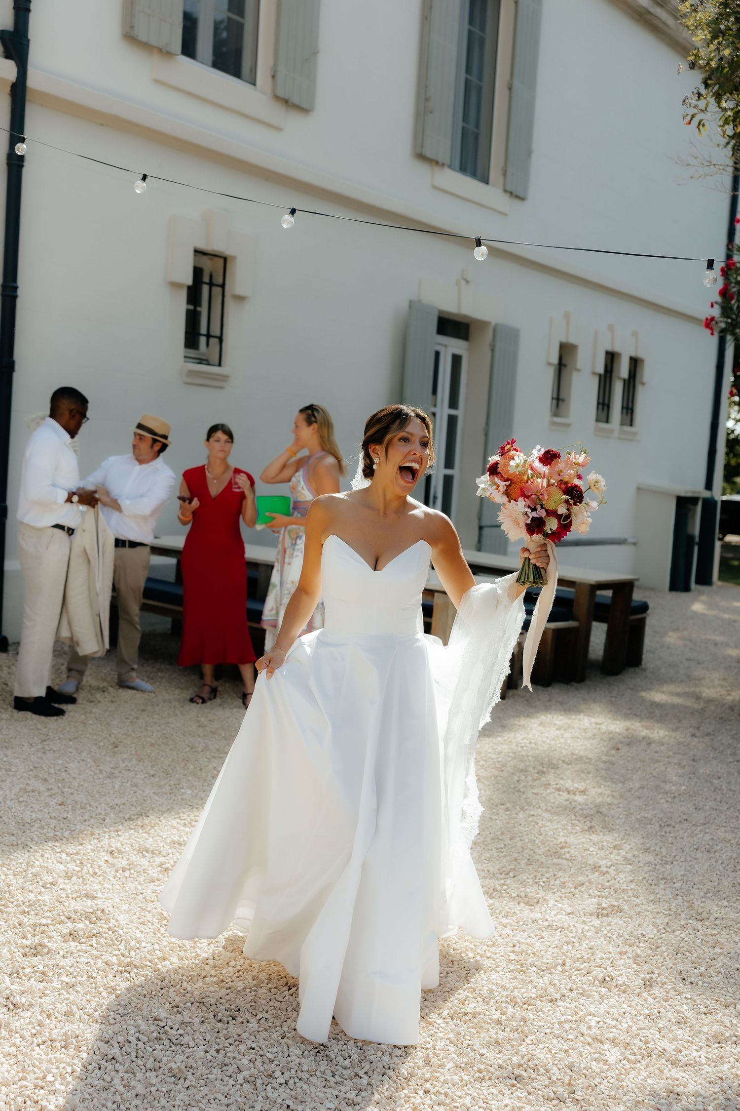 Wedding party in white dresses celebrating outside a white building on sunny day.