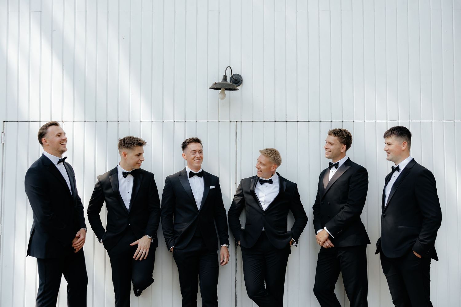 A group of groomsmen in black tuxedos stand together in a row against a light colored wall.
