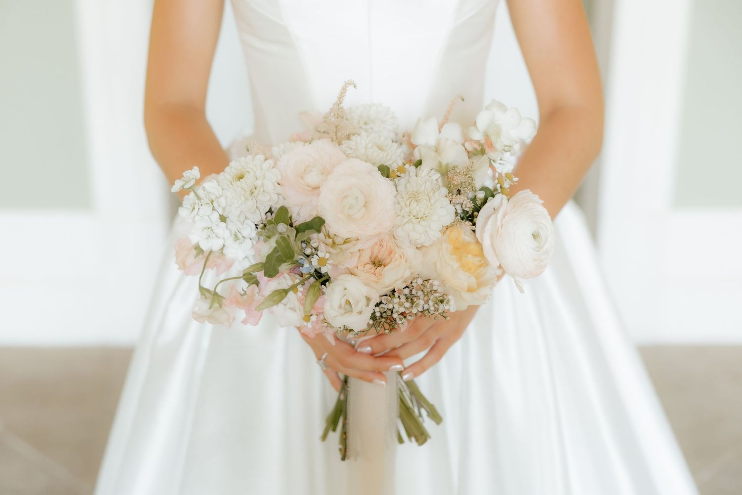A bride in a white wedding gown holds a delicate bouquet of pink and white flowers against a bright background.