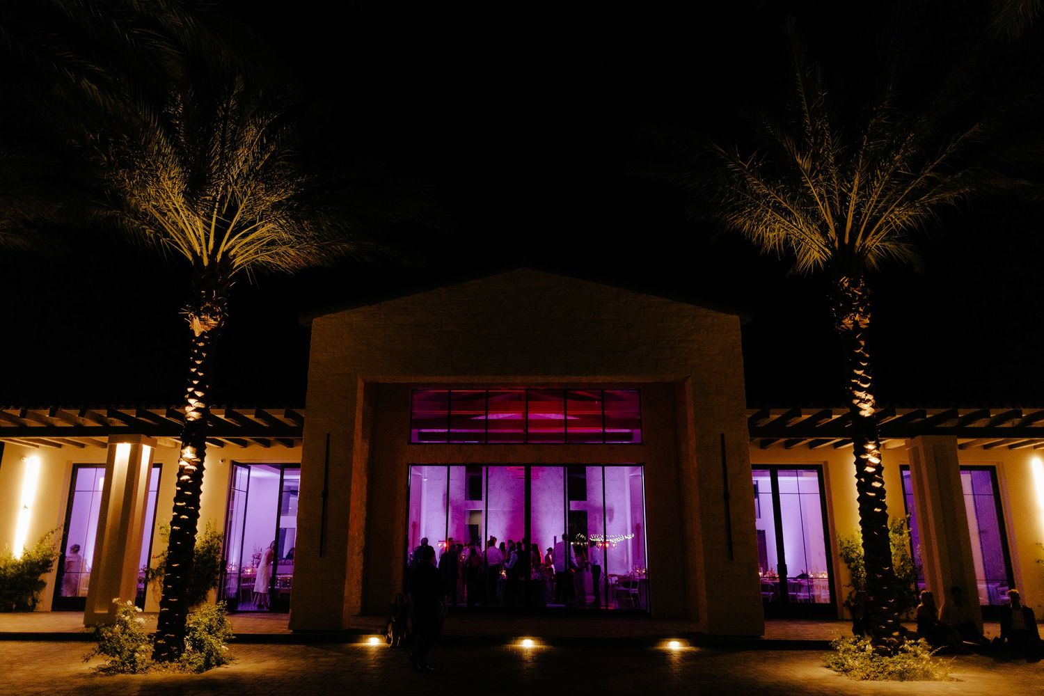 Stunning wedding venue exterior at night with purple uplighting and illuminated palm trees flanking entrance.