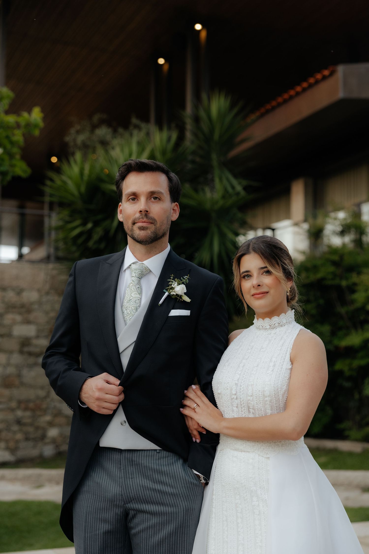 Elegant bride and groom pose together outdoors in front of modern building with tropical greenery.