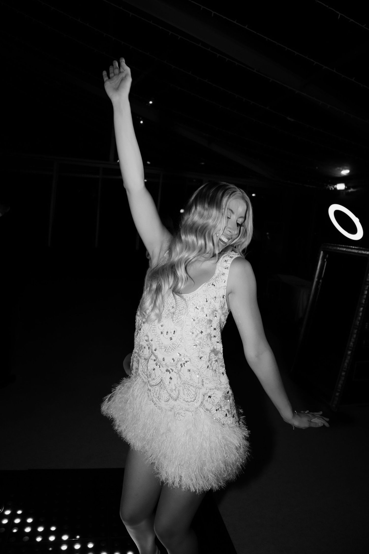 Bride dancing joyfully in a white feathered mini dress at her wedding reception in black and white.
