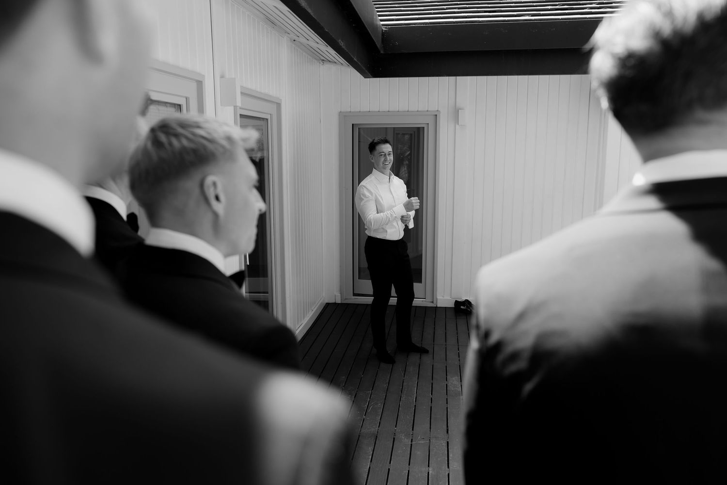 Black and white photo of groom in white shirt standing in a doorway as groomsmen watch during wedding preparations.