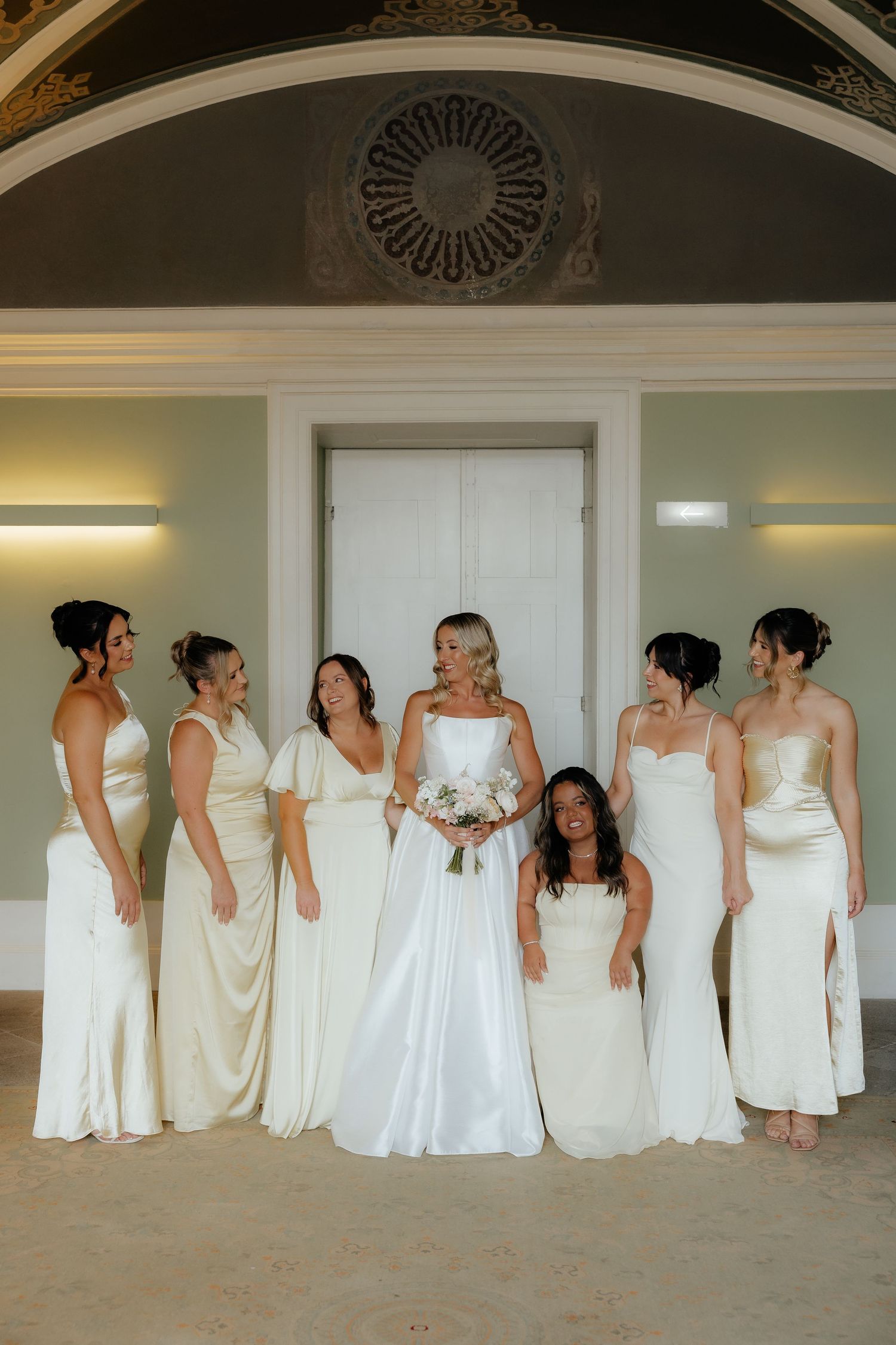 Bride in white satin gown poses with six bridesmaids in ivory dresses inside elegant venue.