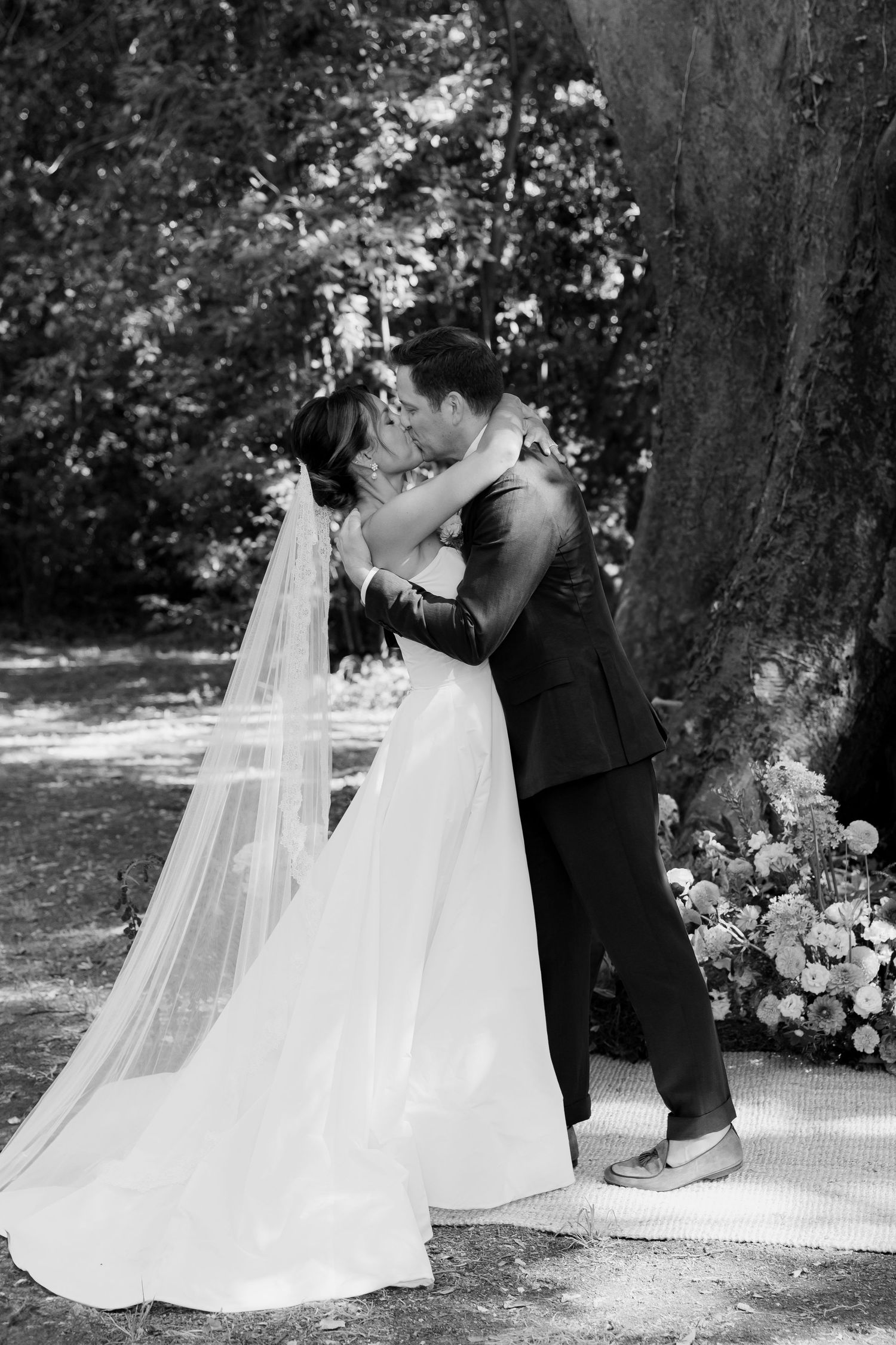 An intimate wedding moment photographed in black and white under a garden tree.