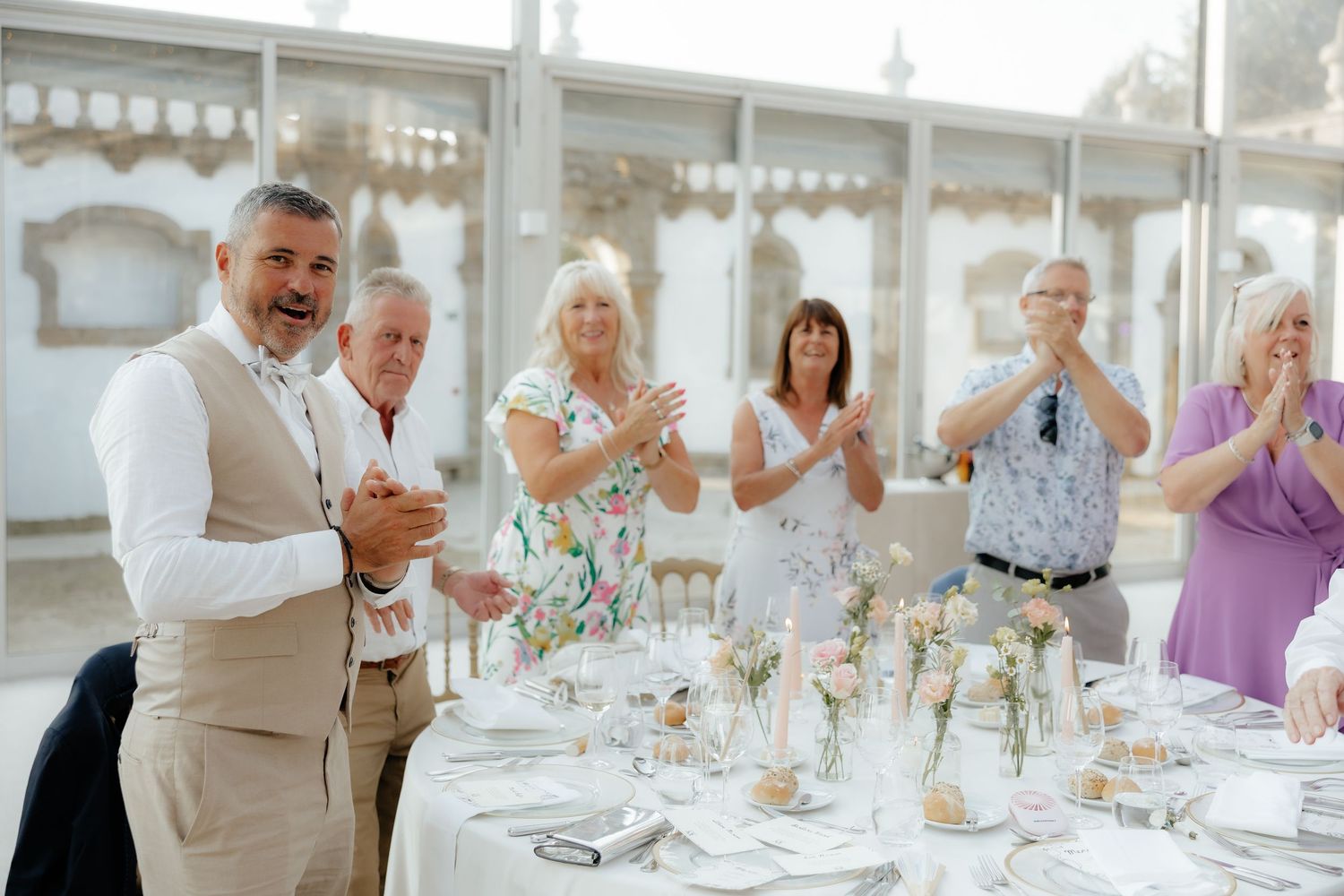 Wedding guests at a reception raise their glasses in a celebratory toast at an elegant white-themed venue.